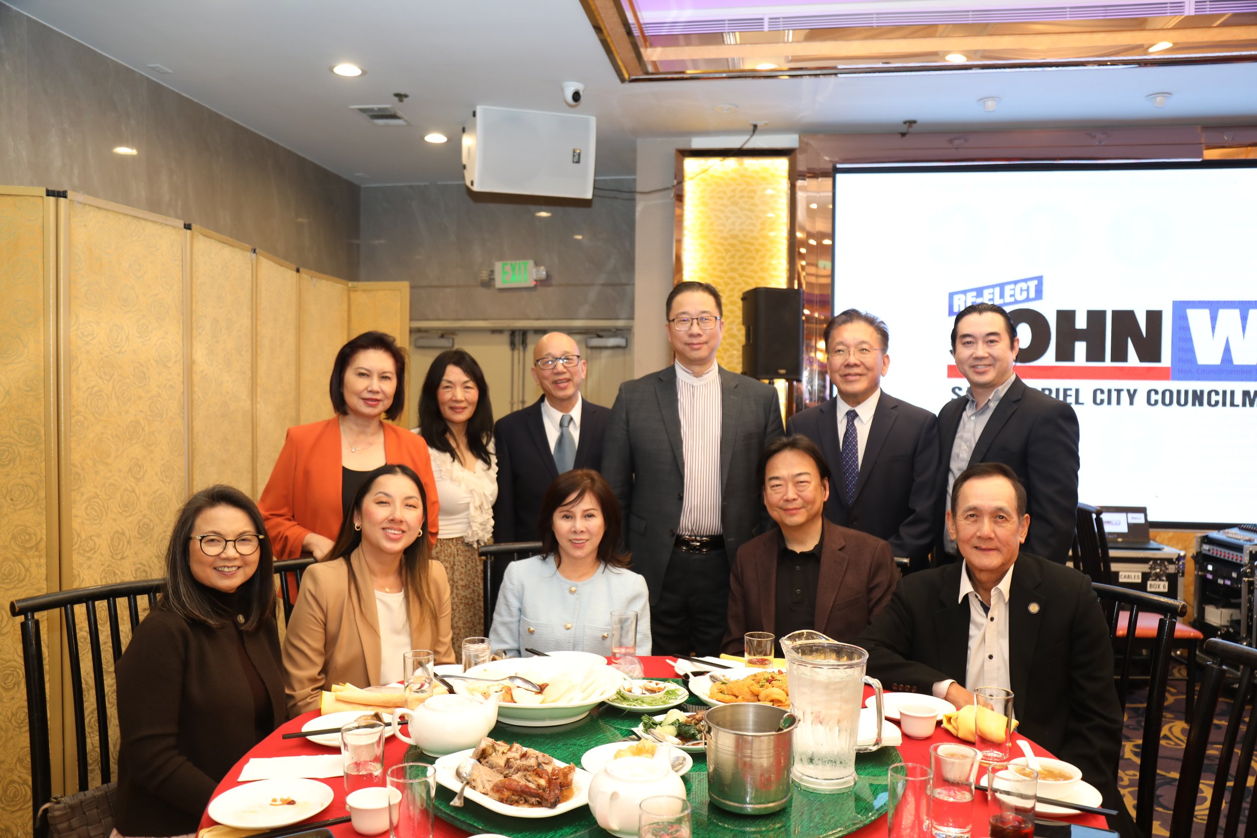Group of diverse people at a formal event or banquet, standing and sitting around a table with food and drinks, with a large screen and decorative lighting in the background.