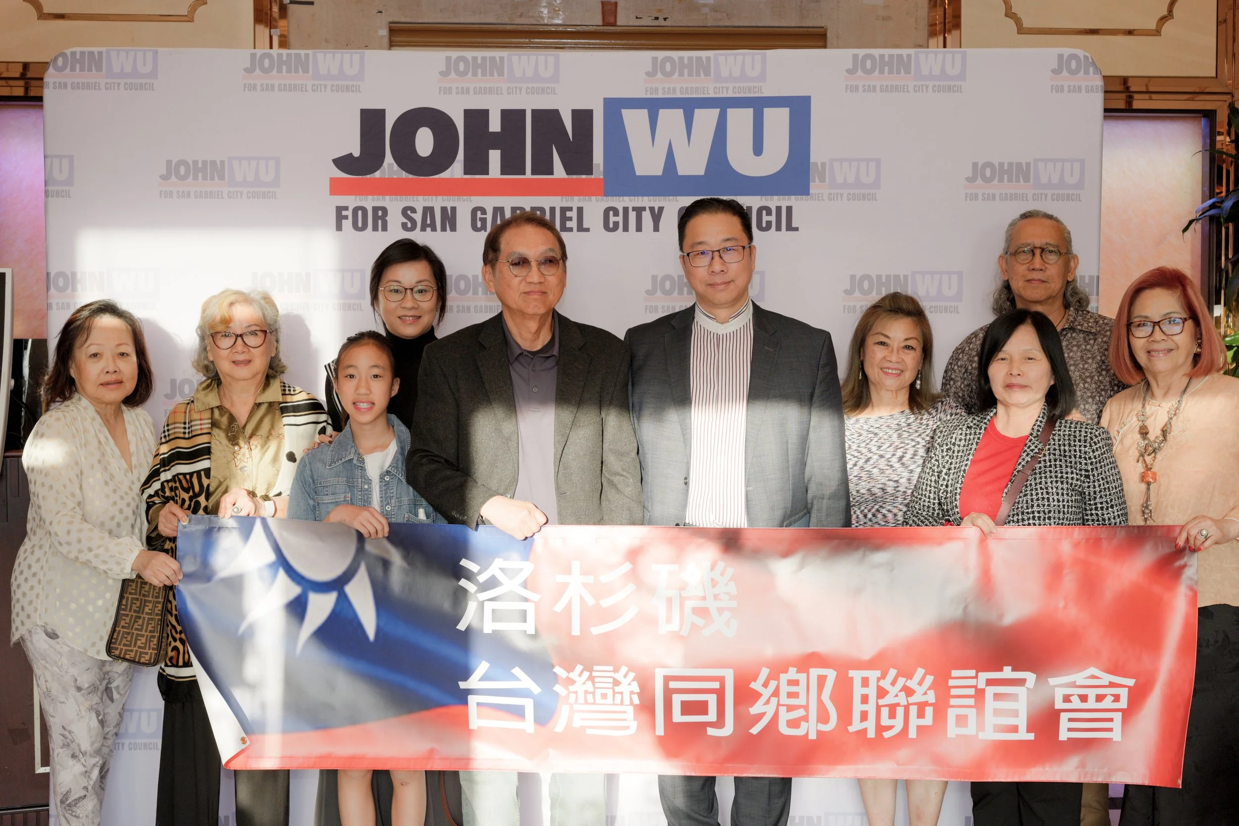 Group of people standing together in front of a campaign banner for John Wu for San Gabriel City Council. The group includes men, women, and a young girl, all smiling and posing for the photo.
