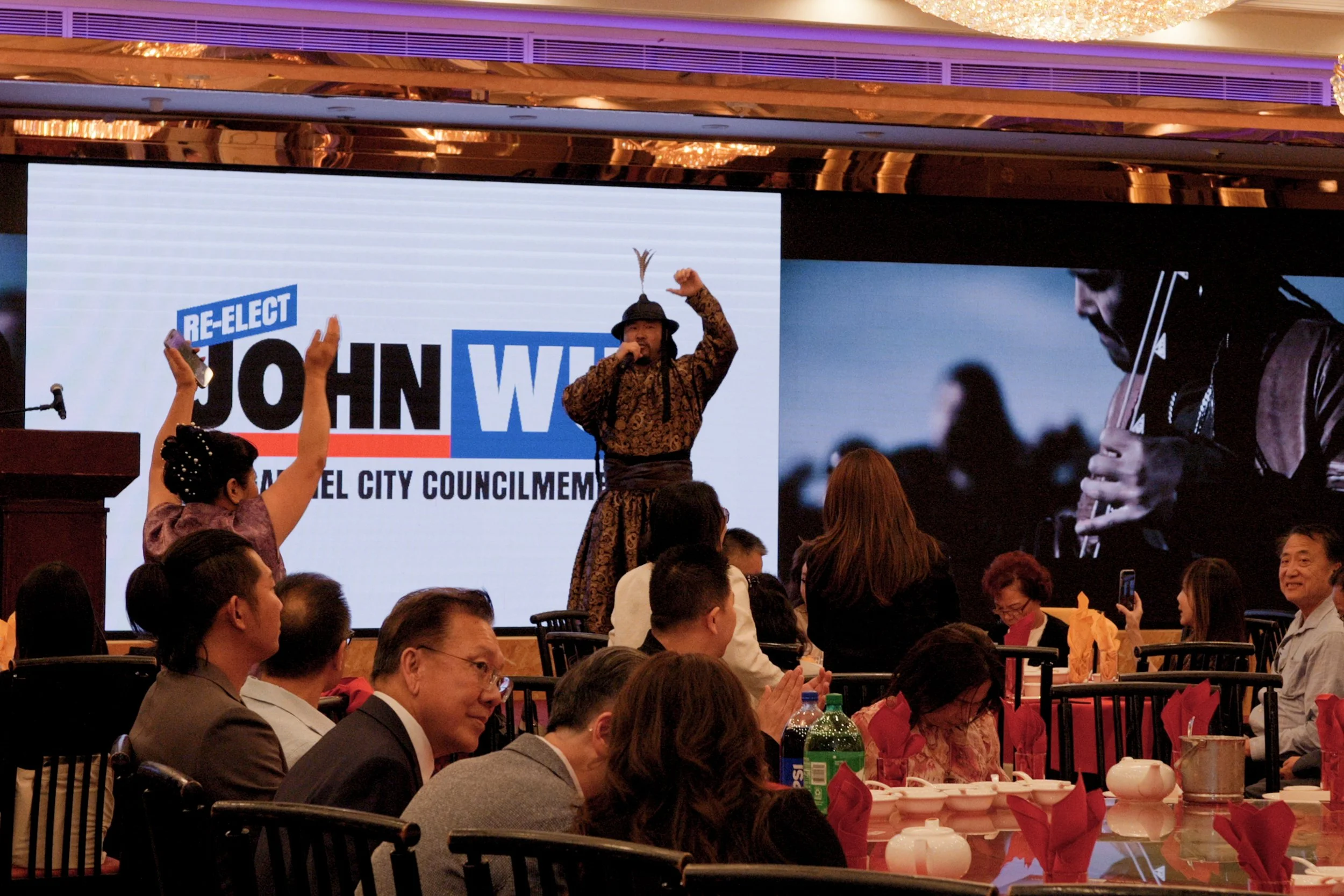 A man in traditional attire speaking on stage at a political event, with a background screen displaying a campaign message supporting John W. for the council. Audience members are seated at tables, some raising their hands.