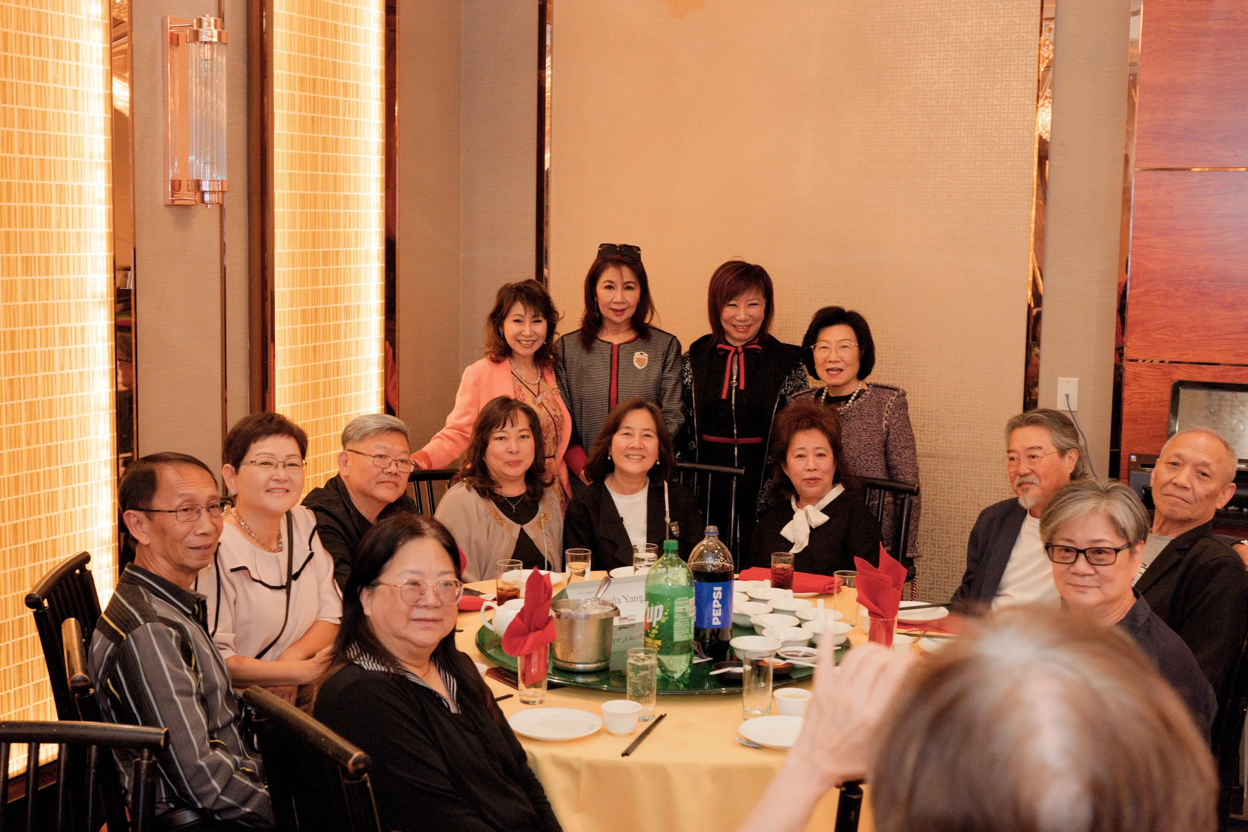 A group of Asian and Caucasian adults gathered at a banquet table in a decorated banquet hall, smiling for a photo during a social event.