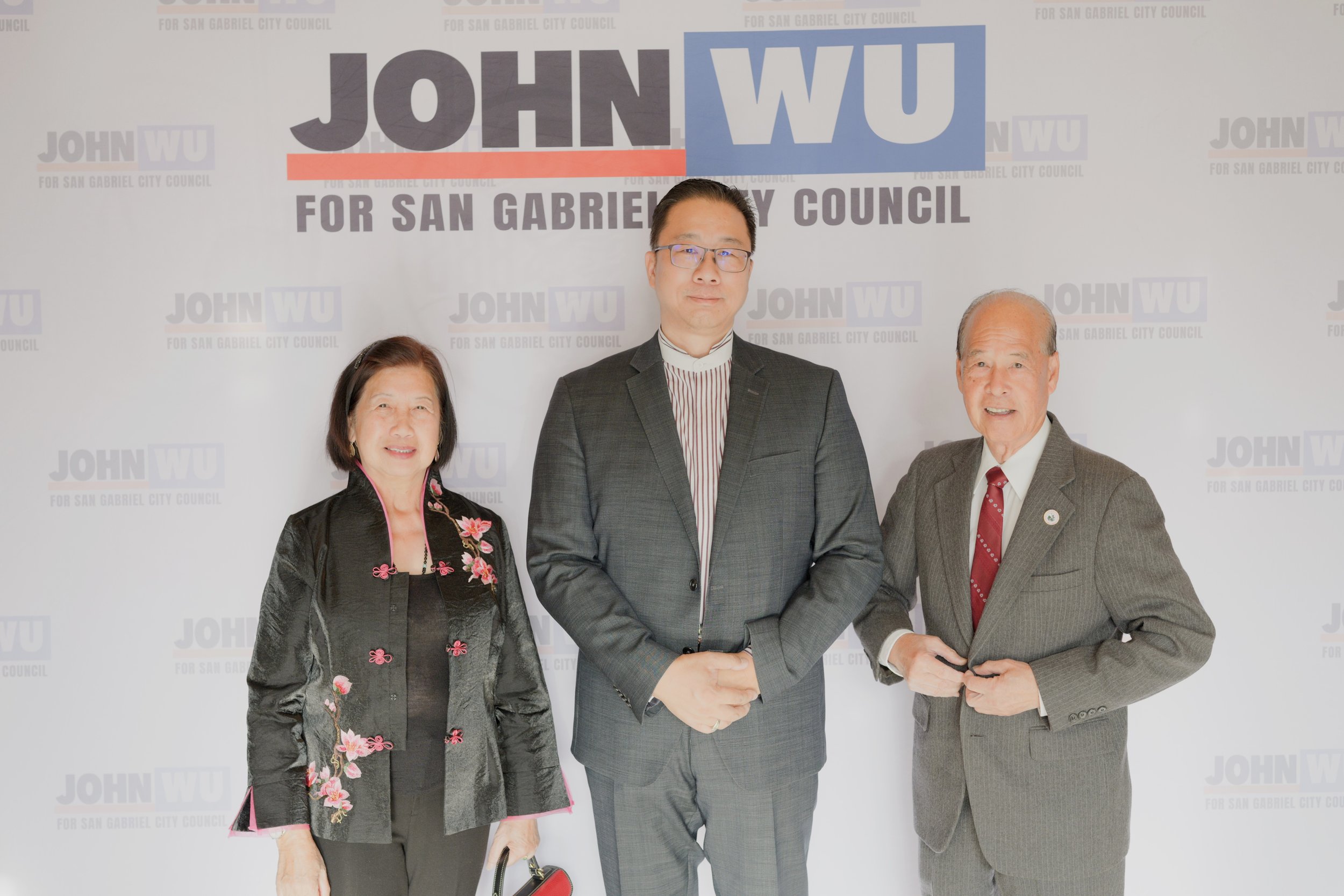 Three people standing in front of a backdrop with text that reads "John Wu for San Gabriel City Council." The woman on the left is wearing a black silk jacket with pink and floral embroidery, the man in the middle is in a gray suit with a striped shi