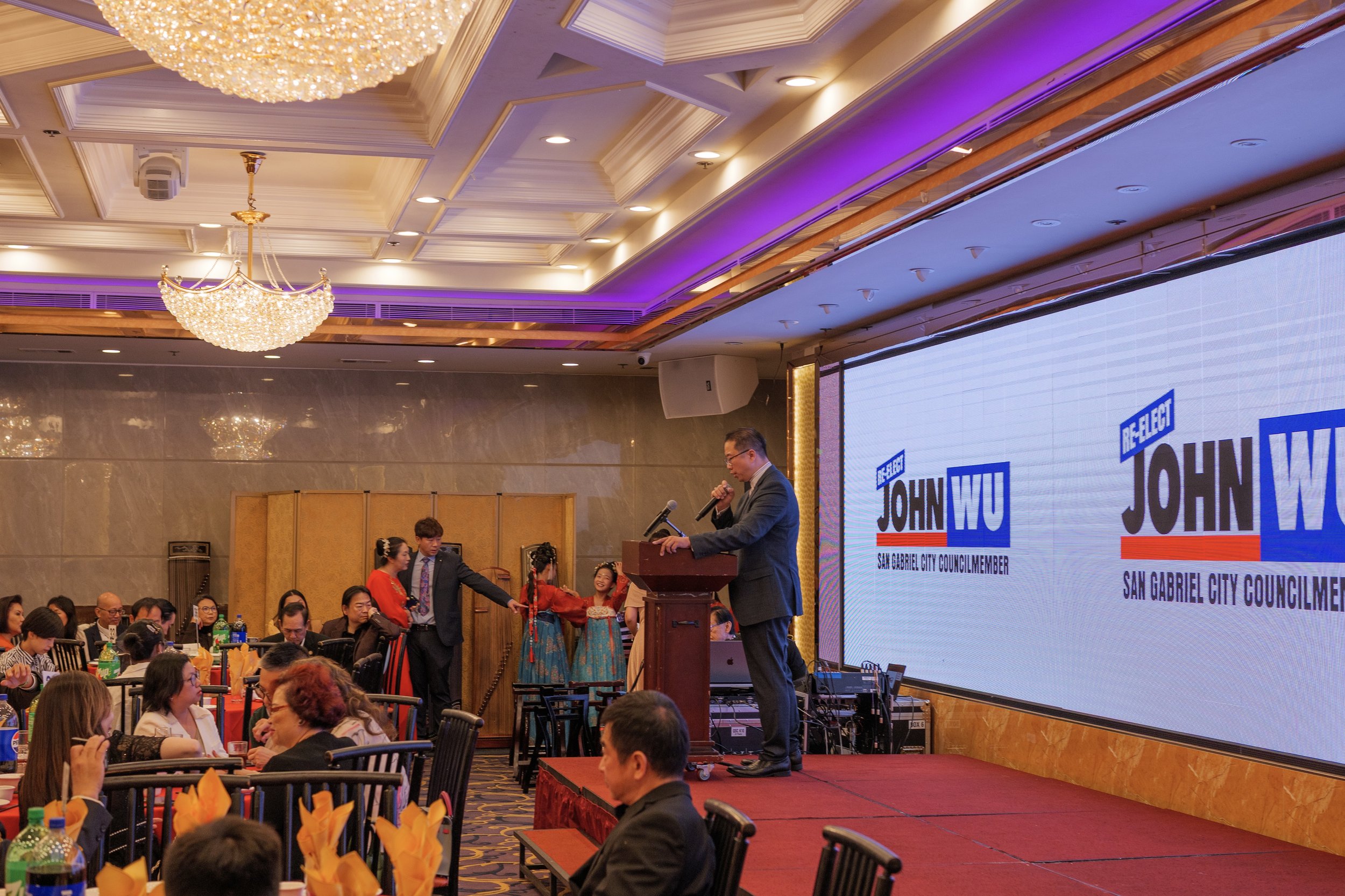 A man in a suit speaking at a podium during a political event in a banquet hall, with a digital screen displaying 'John W.' and 'San Gabriel City Council Member' behind him. Several dancers in traditional attire are on stage, performing for the audie