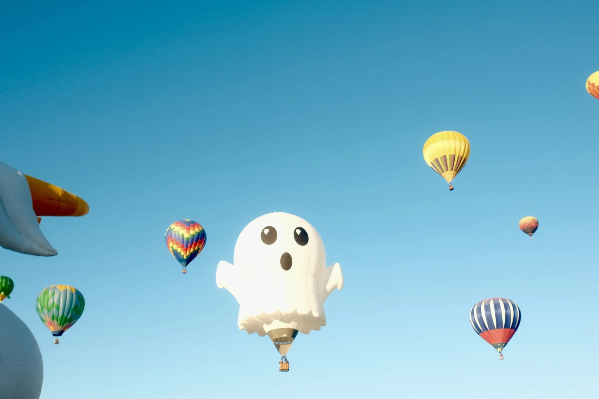 A ghost-shaped special shape balloon floating among other colorful balloons in a clear blue sky