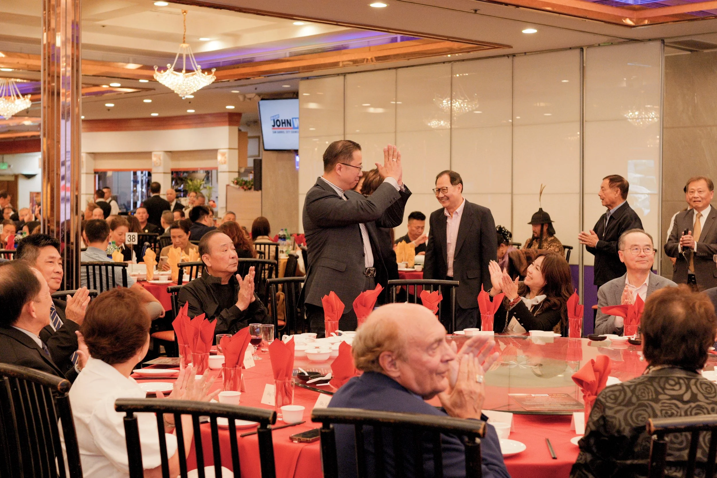 People gathered at a banquet or formal event in a decorated hall with chandeliers, tables with red cloths and napkins, and a man in a dark suit receiving applause.