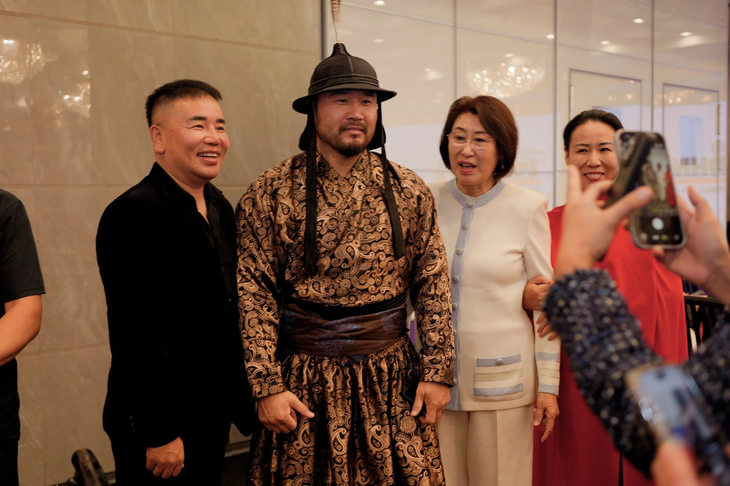 Group of four people taking a photo together in an indoor setting, with one person wearing traditional Asian attire and a hat, while the others are dressed in modern clothing.