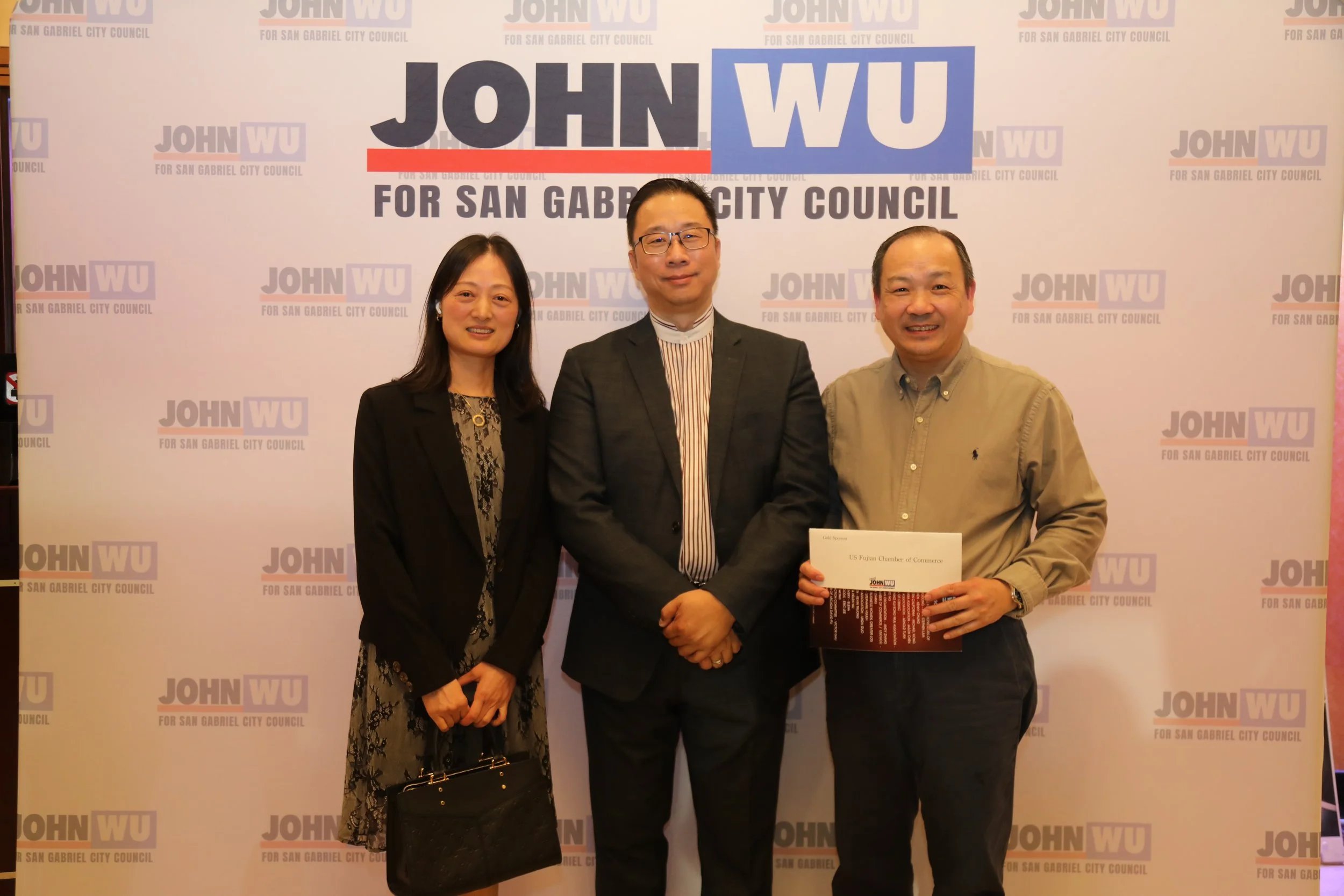 Three people standing together in front of a backdrop with campaign logos. The person on the right holds a certificate. The backdrop shows the name 'John Wu' and mentions the San Gabriel City Council.