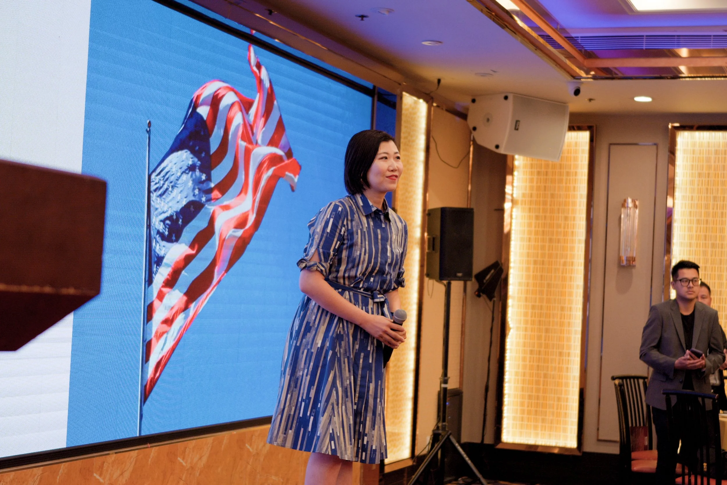 A woman in a blue patterned dress holding a microphone standing in front of a large screen displaying an American flag.