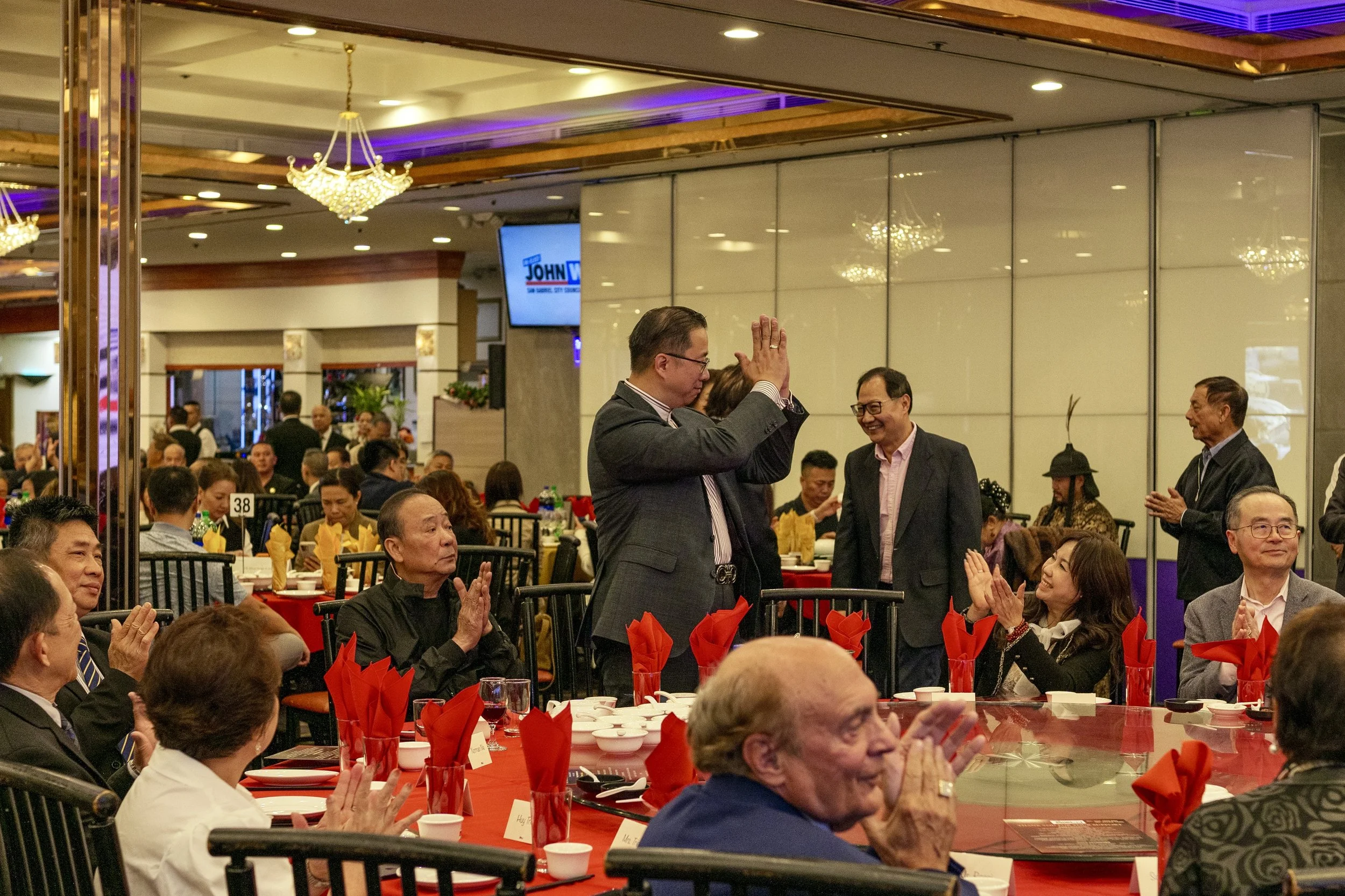 A man in a suit is standing with his hands clasped in front of his face, smiling at a seated woman during a formal event in a decorated banquet hall filled with people at round tables.