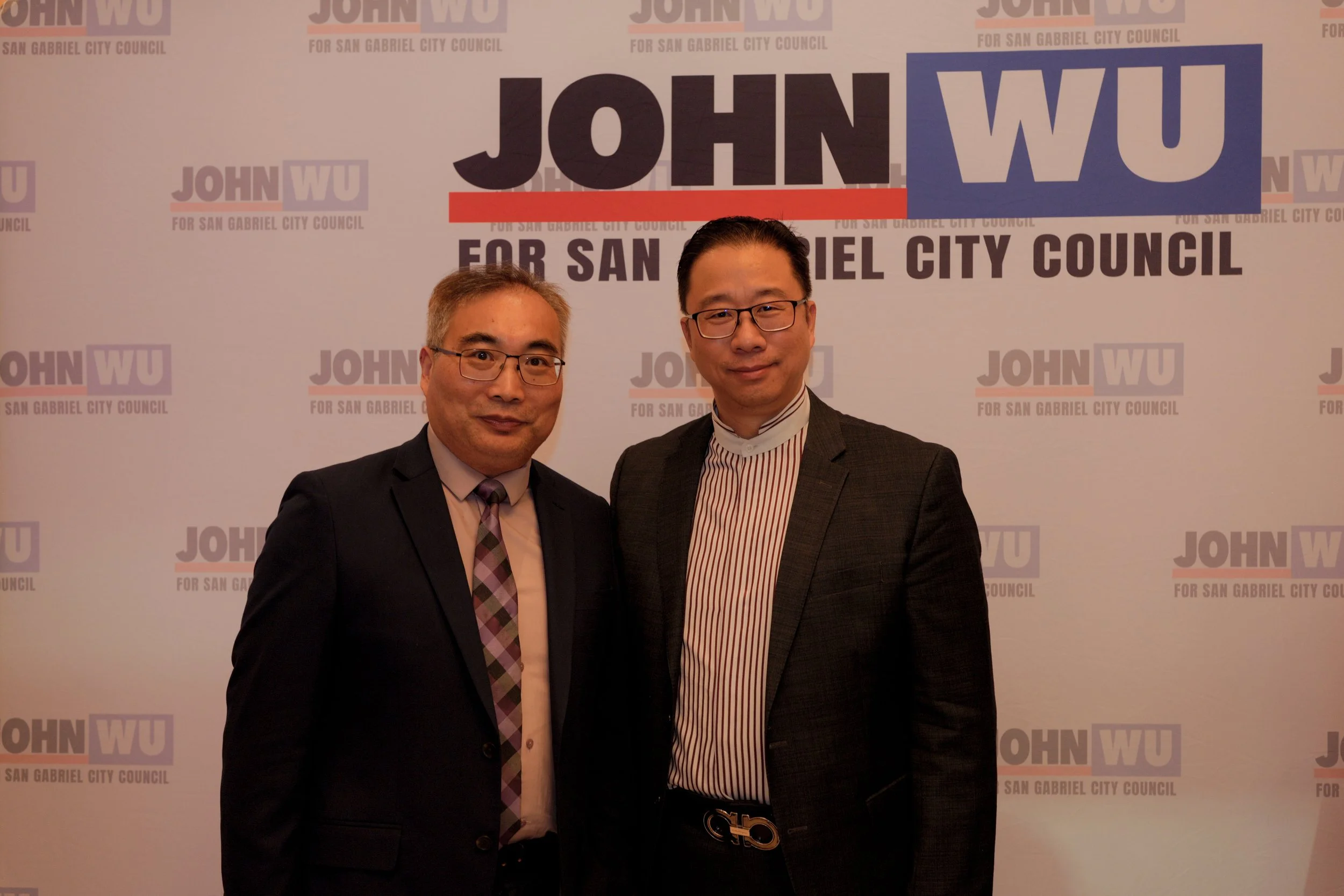 Two men in suits standing in front of a backdrop with the text "John Wu for San Gabriel City Council." both men are smiling, one has glasses, and the backdrop repeats the campaign slogan.