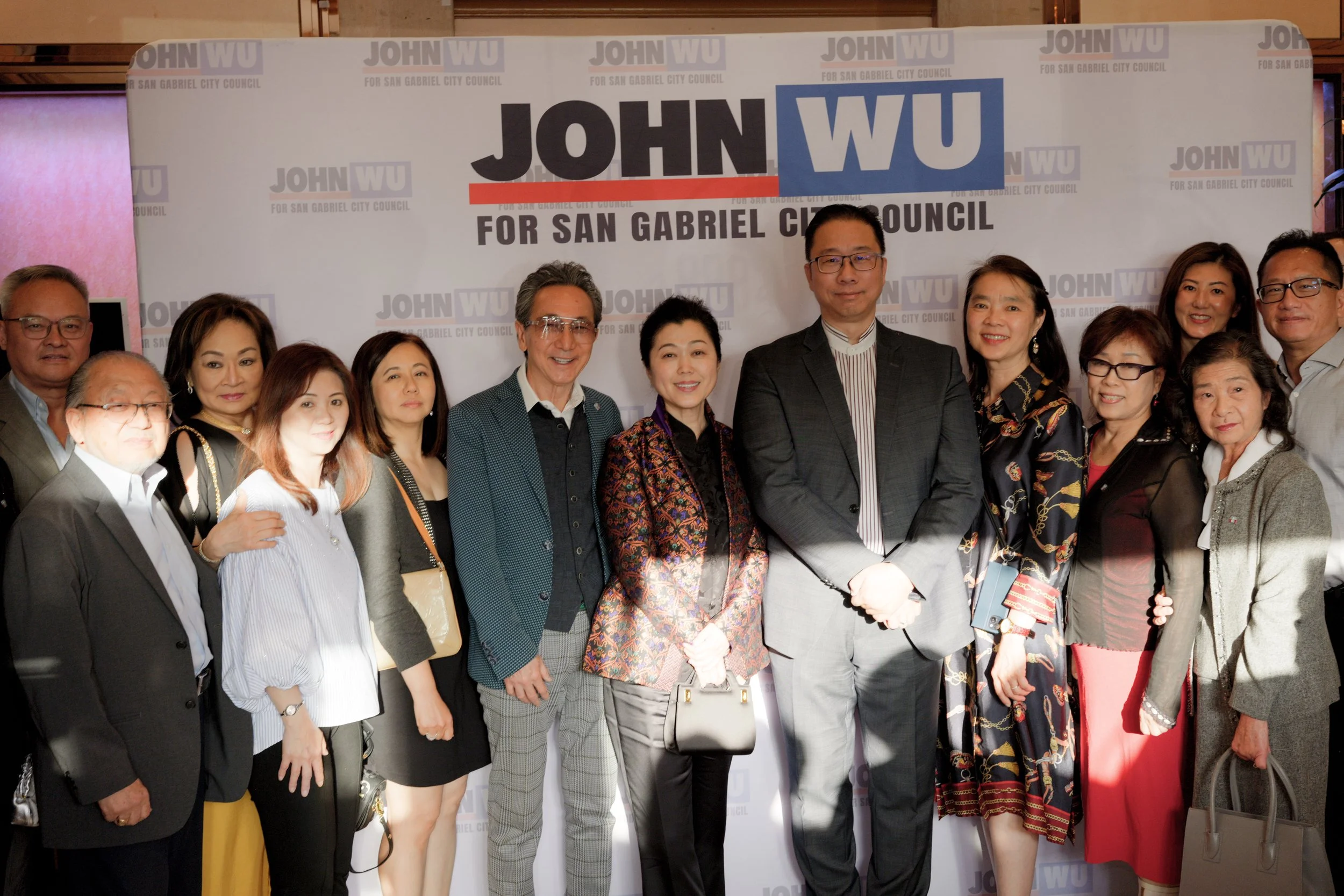 Group of people standing in front of a campaign banner that reads "JOHN WU FOR SAN GABRIEL CITY COUNCIL", smiling for the photo.
