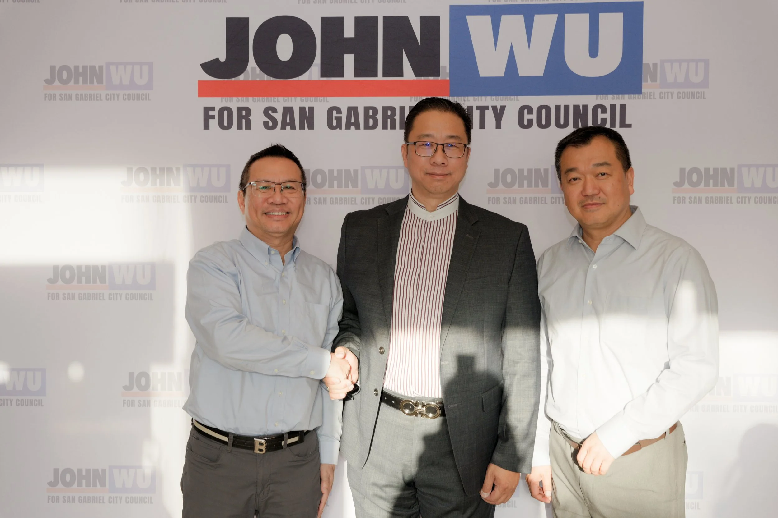 Three men are standing together in front of a backdrop that reads "JOHN WU for San Gabriel City Council." The man in the middle is shaking hands with the man on the left, both of whom are smiling, while the man on the right looks on. All three men ar