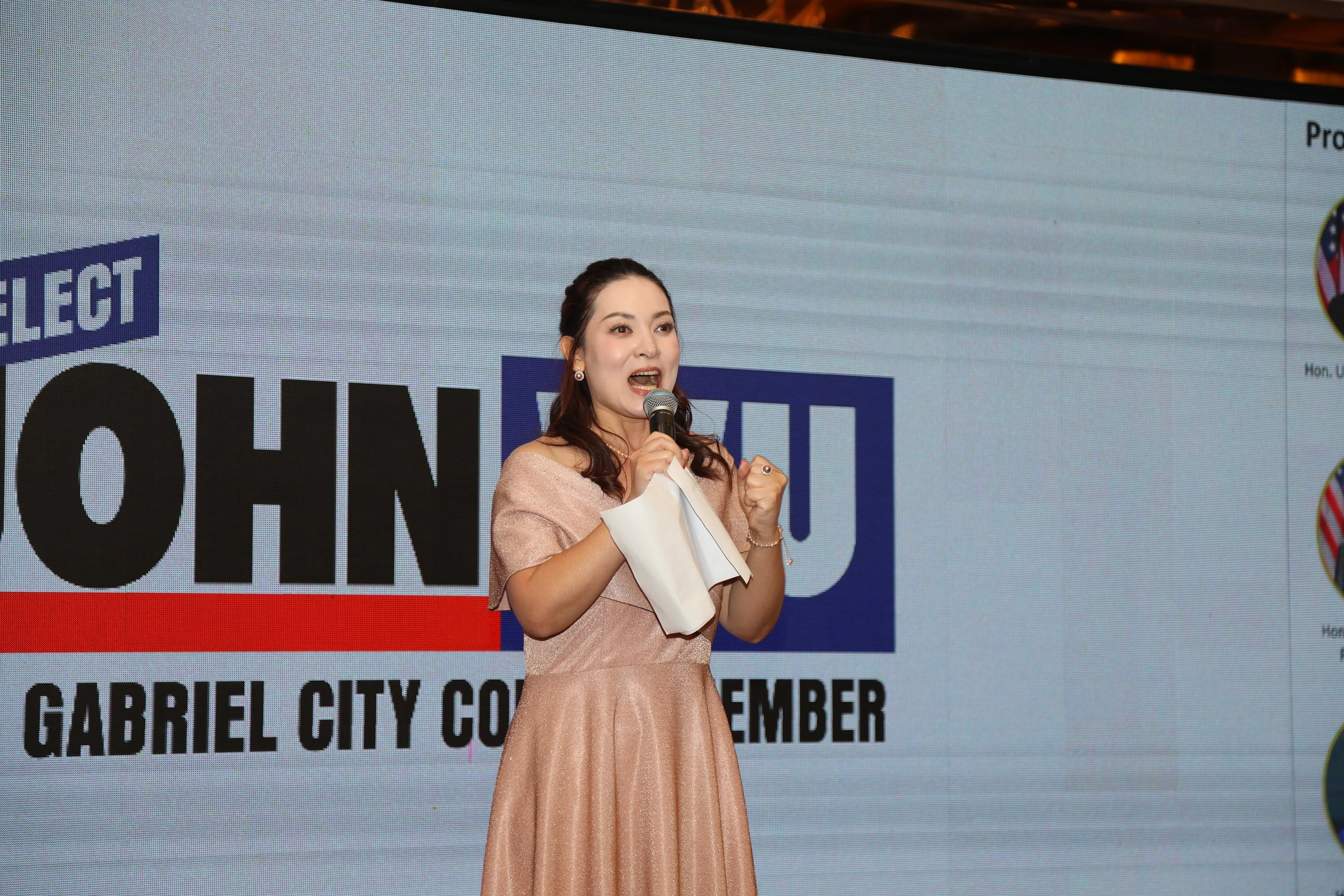 A woman in a pink dress speaking into a microphone on stage during an event in Gabriel City, Philippines, with a large screen displaying a political campaign sign behind her.
