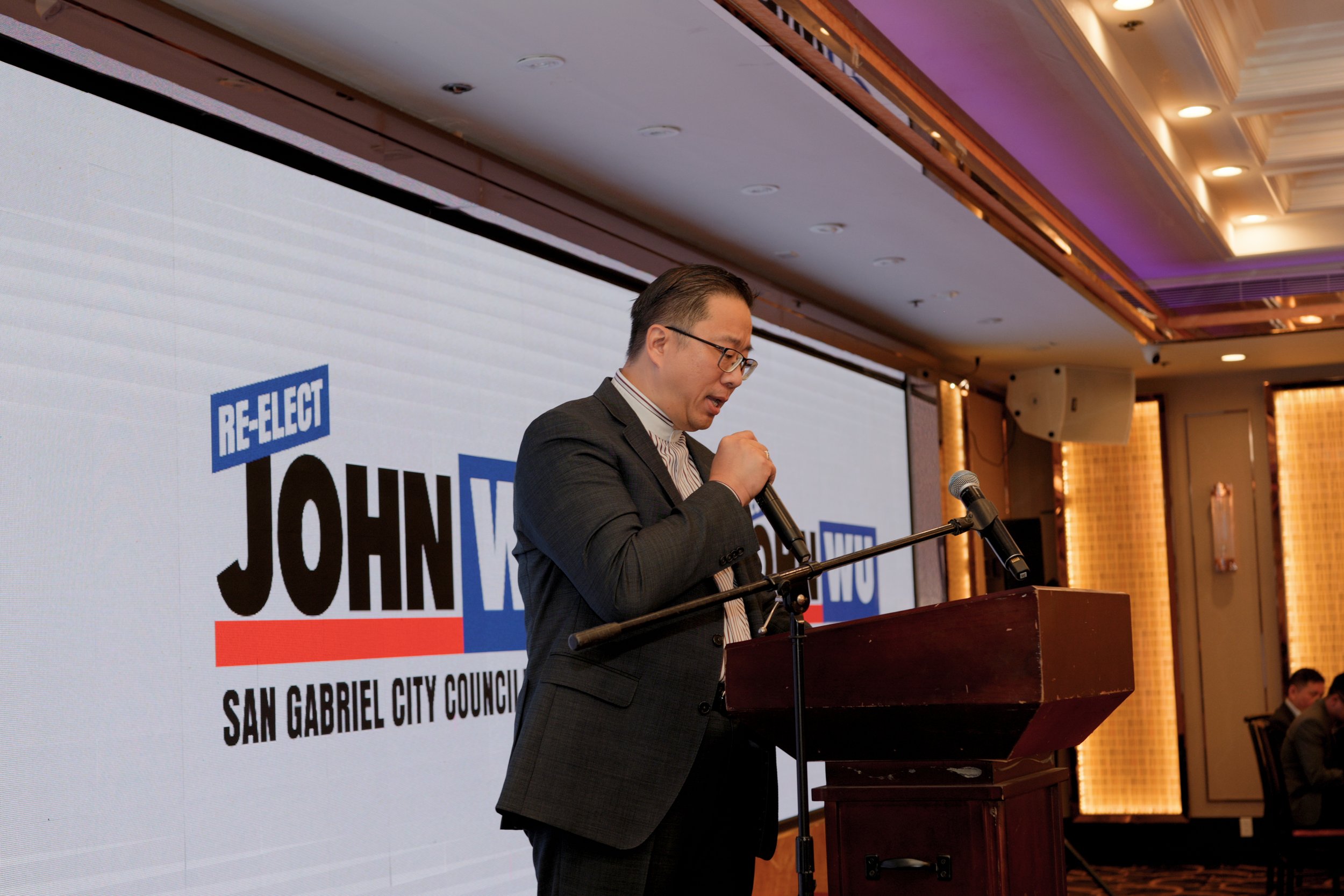 A man in a suit and glasses stands behind a podium during a formal event, with a large screen behind him displaying political campaign signs promoting John V from San Gabriel City Council.