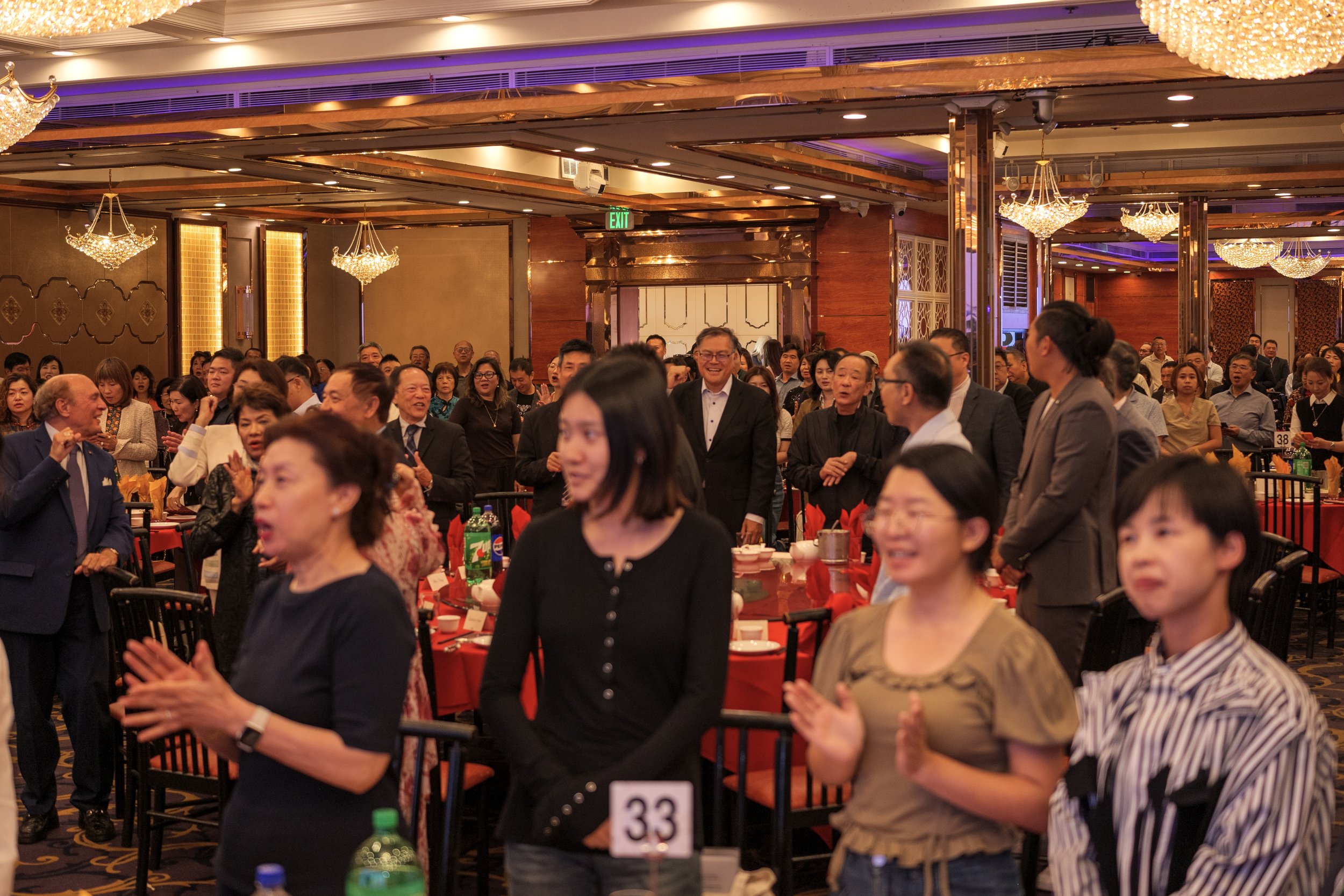 Crowd of diverse people in formal attire gathered in a banquet hall with chandeliers, seated at round tables covered with red tablecloths, participating in an event or celebration.
