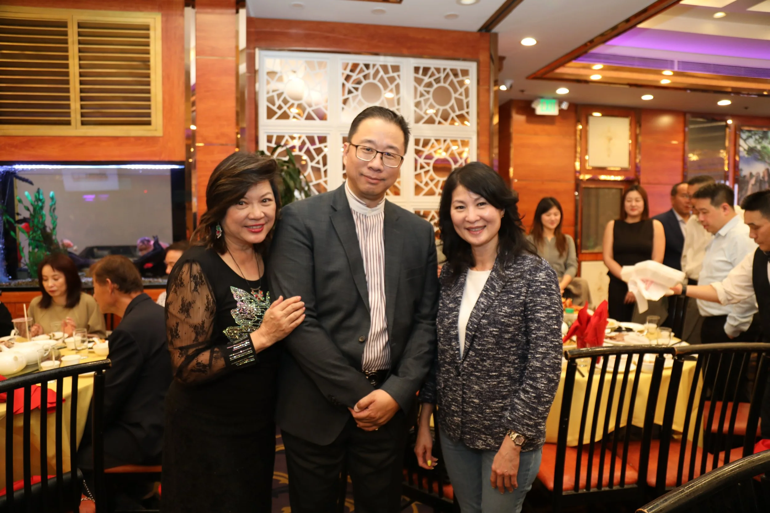 Three people standing and smiling at a social event, with a group of people and decorations in the background.