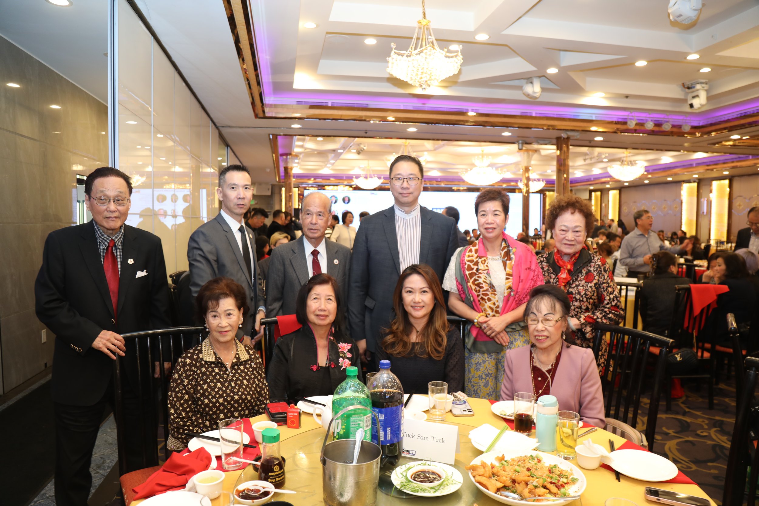 A group of Asian men and women, some standing, posing for a photo at a formal gathering or celebration in a banquet hall with purple lighting and chandeliers. The table in front has food and drinks, including dishes, bottles, and glasses.