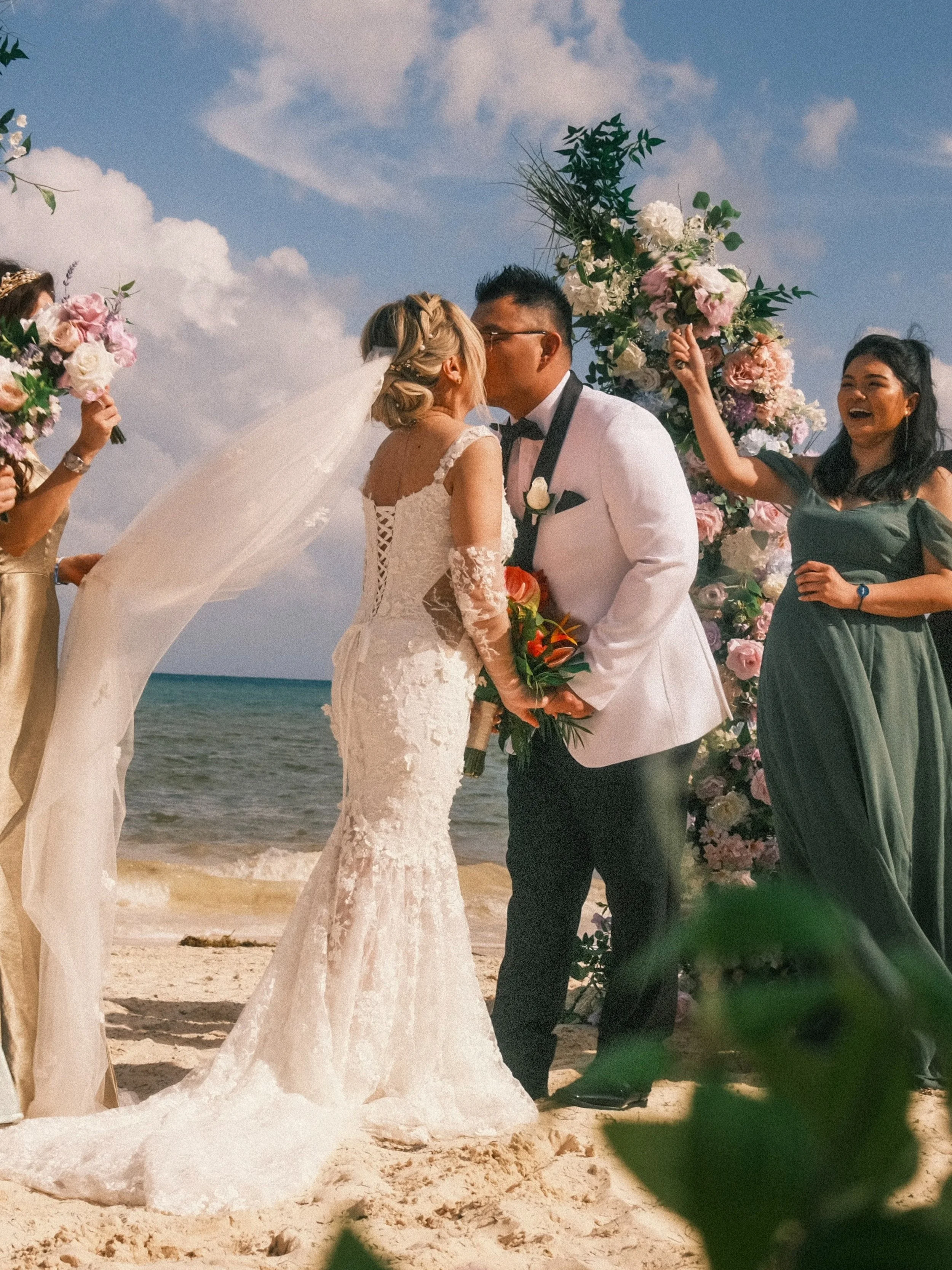 A couple kissing during their beach wedding ceremony on the sand, with a floral arch and bridesmaids holding bouquets nearby.