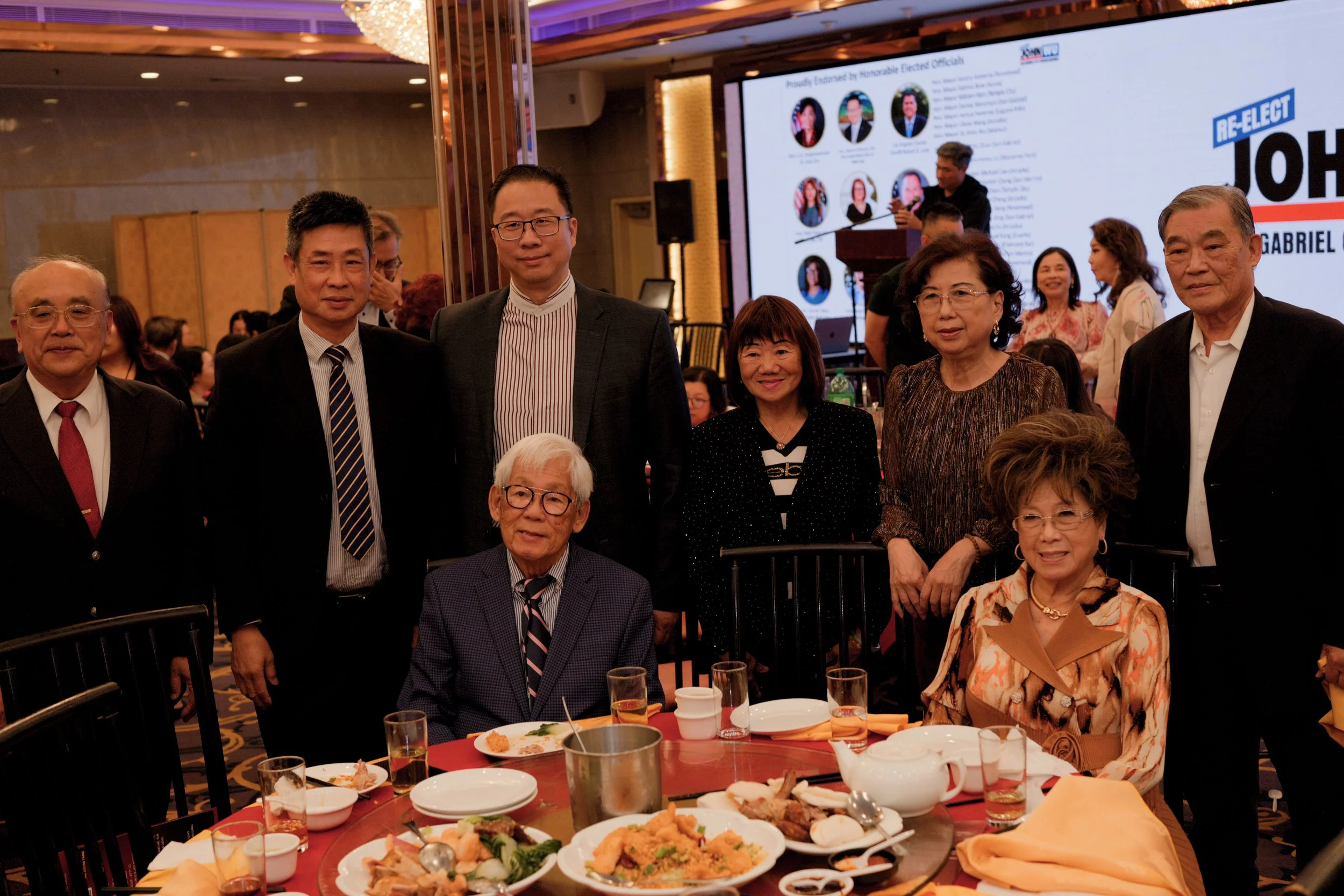 Group of diverse people at a formal event or banquet, with a large screen in the background displaying campaign graphics for re-election of a candidate named John Gabriel, and a table in front with partially eaten food and drinks.