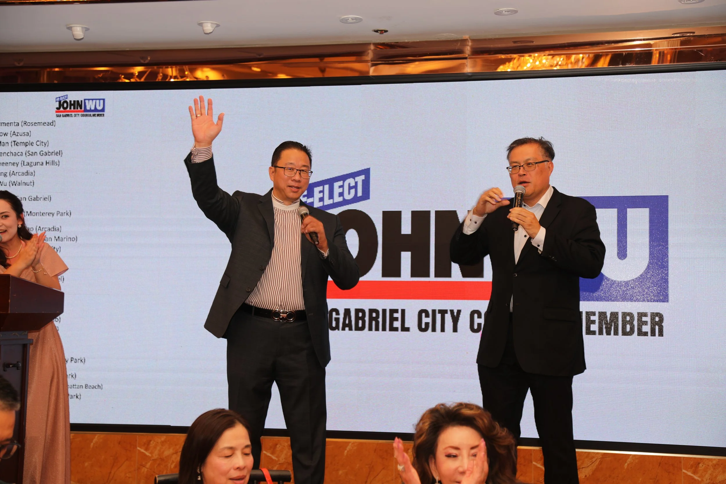 Two men at a political event speaking into microphones, one raising his hand, with a large screen behind them displaying text and the logos of John Wu and the campaign. Several people are seated in front of the stage.
