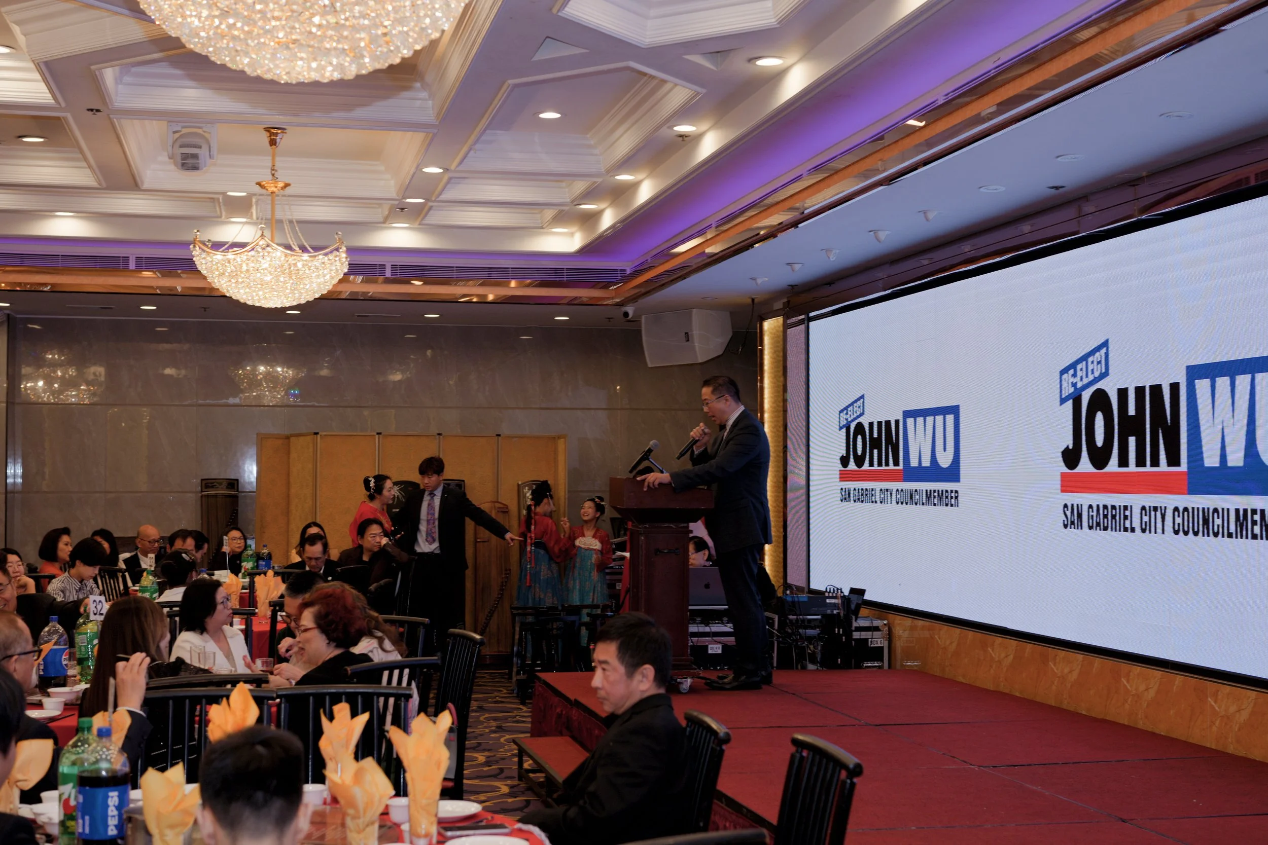 A formal indoor event with a man speaking at a podium, a large screen displaying campaign posters for John W. including the text "San Gabriel City Councilmember," and numerous attendees seated at round tables with orange napkins, surrounded by a deco