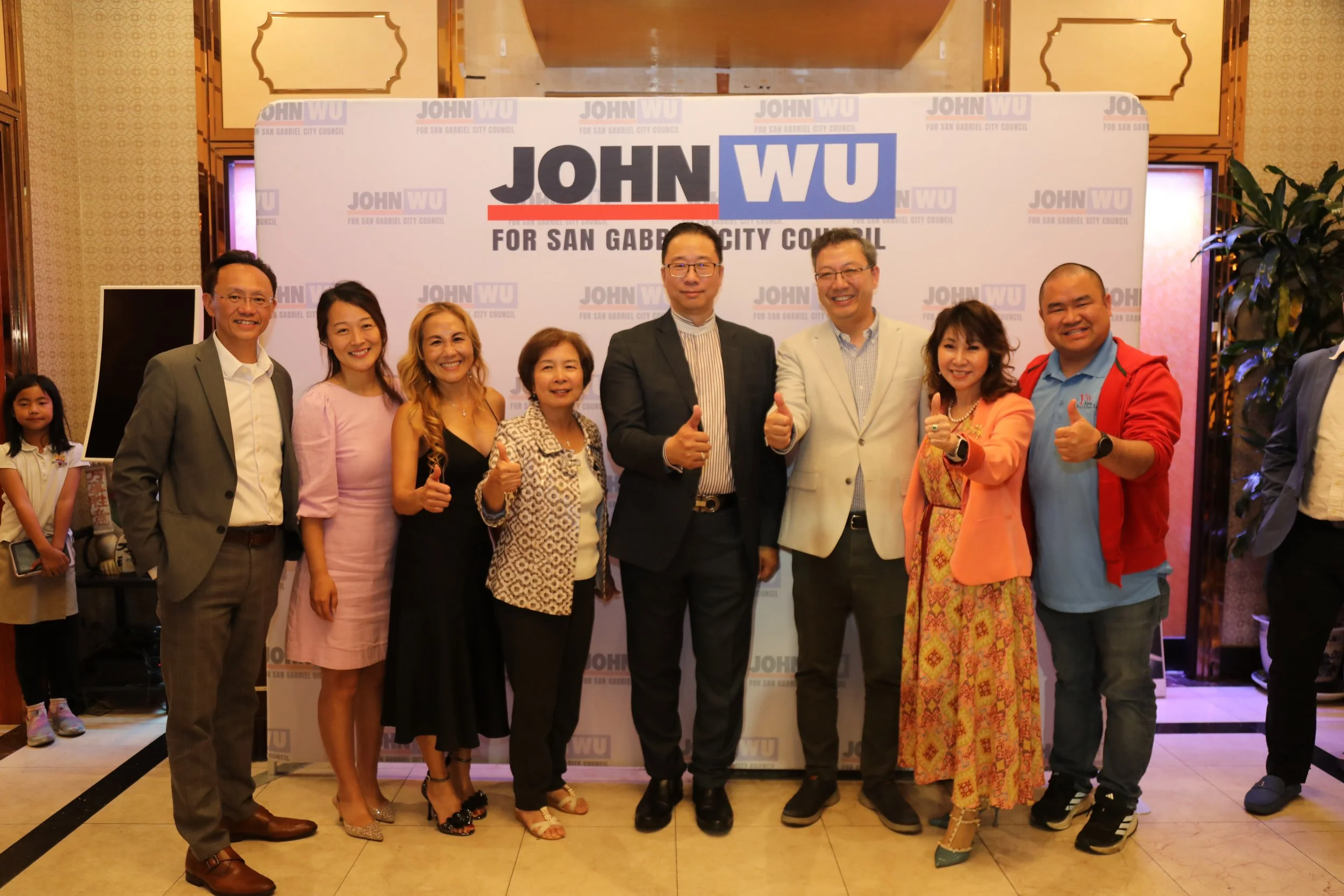 Group of eight people standing in front of a campaign sign for John Wu for San Gabriel City Council, giving thumbs up and smiling.
