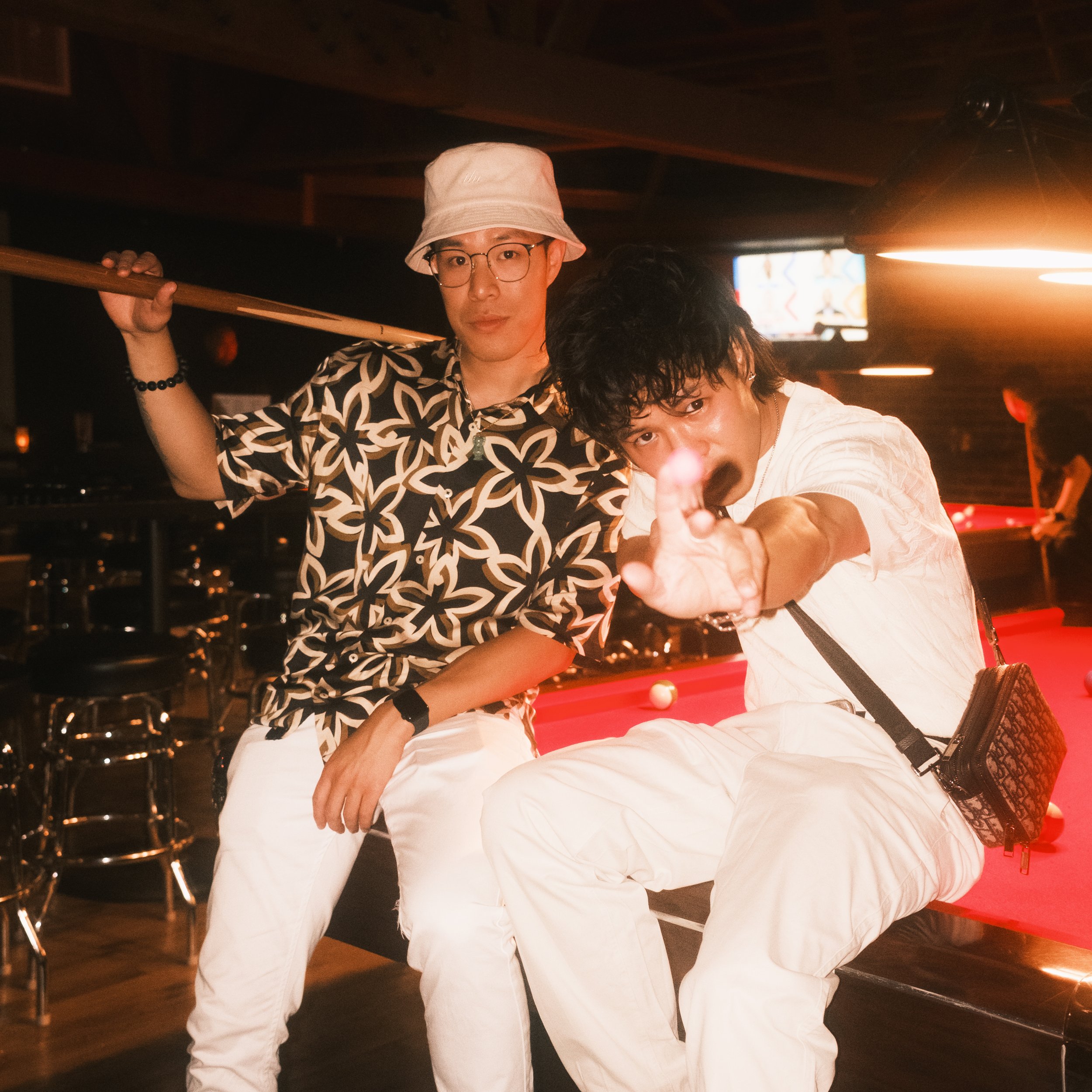 Two young men sitting at a pool table in a bar or club, one wearing glasses and a bucket hat, the other pointing at the camera with a finger, both dressed casually.