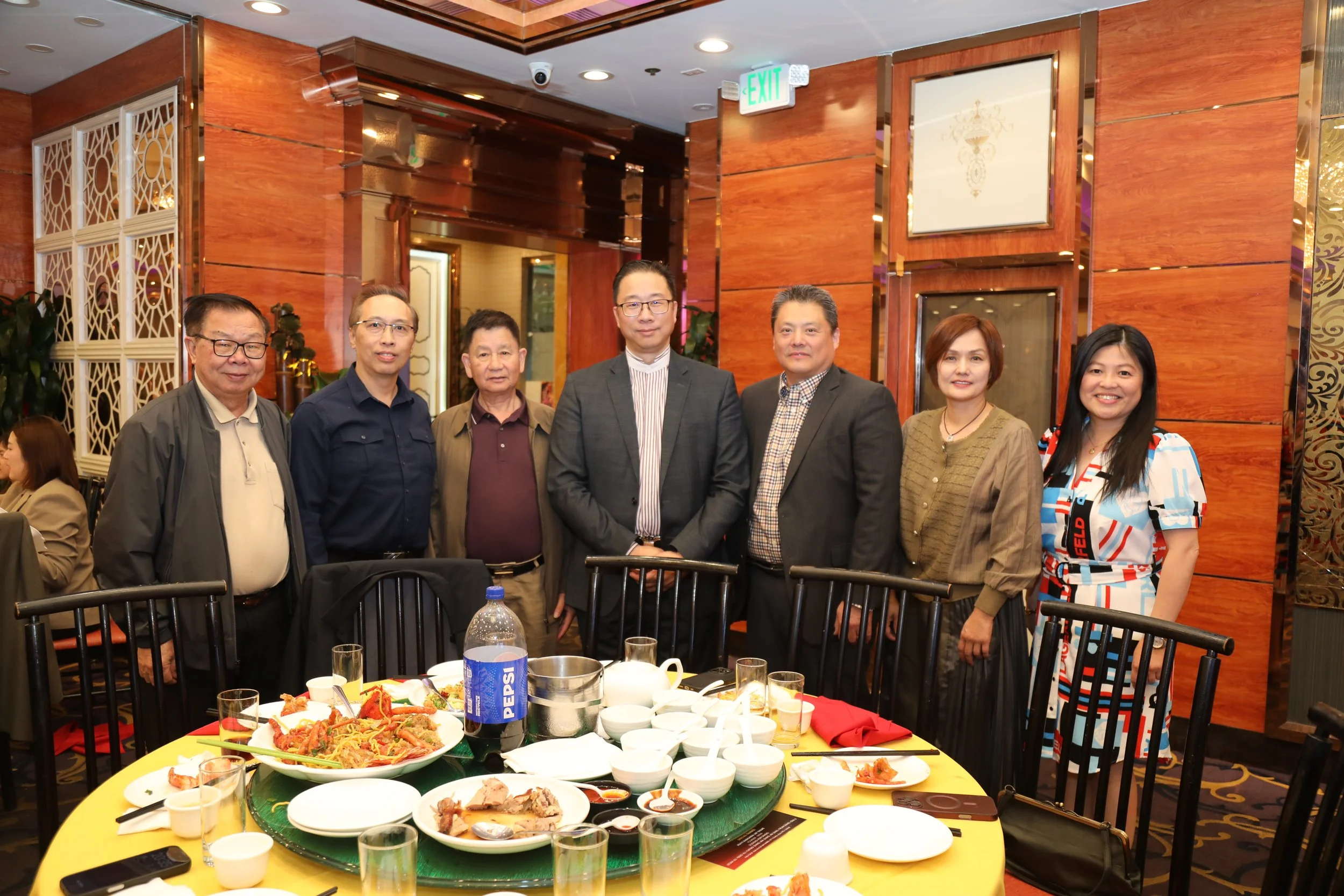 Group of seven people standing behind a table with food in a restaurant or banquet hall.
