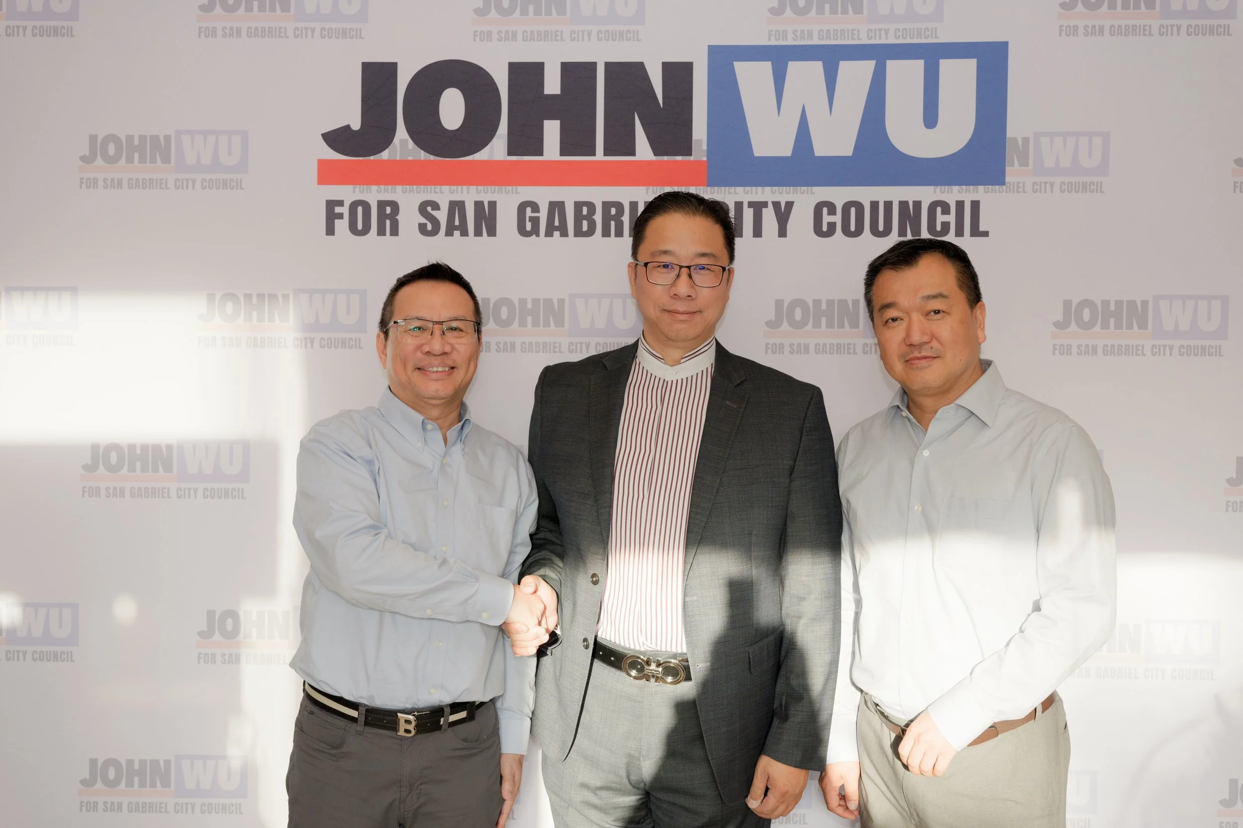 Three men in business attire shaking hands at a political campaign event with a background sign reading "John Wu for San Gabriel City Council."