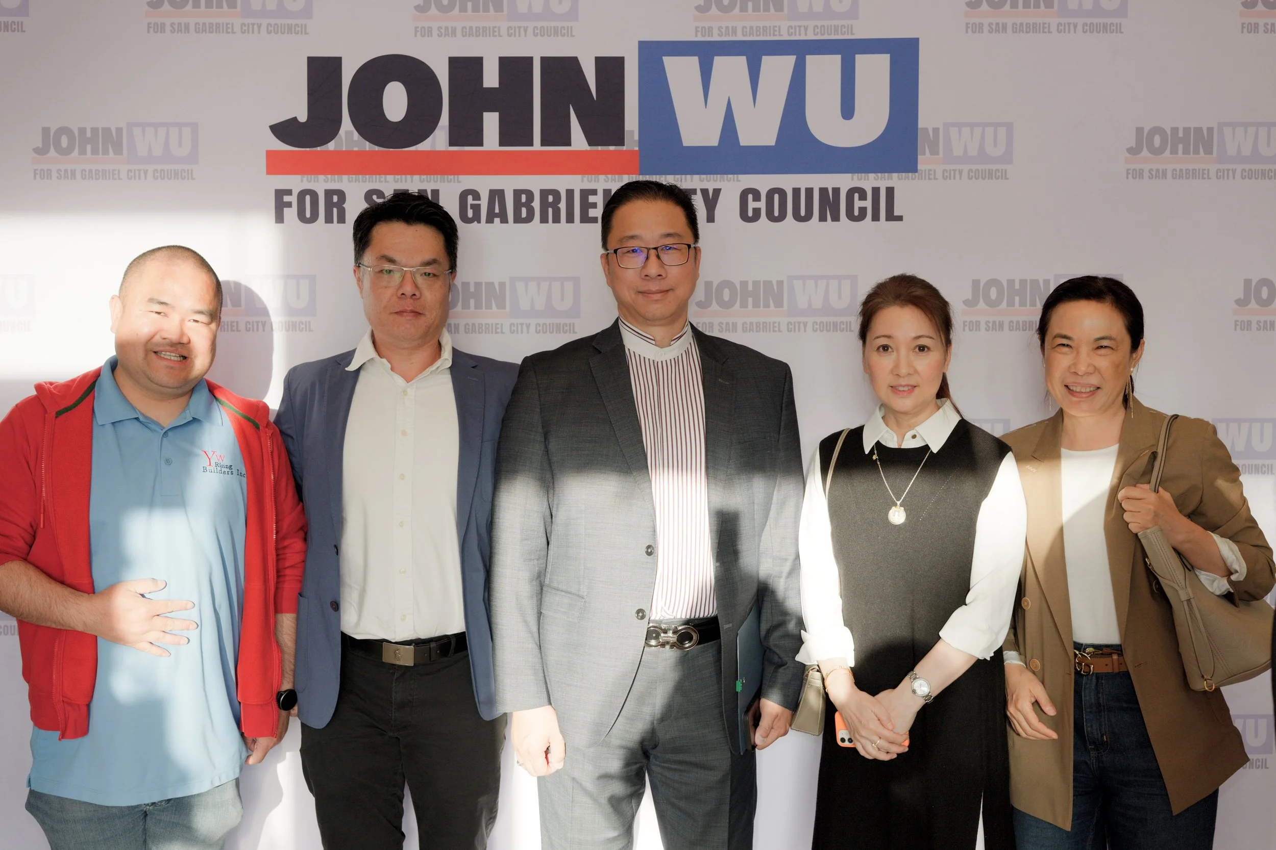 A group of six diverse people standing in front of a campaign backdrop with the words 'John Wu for San Gabriel City Council.' They are dressed in business and casual attire, smiling for the photograph.