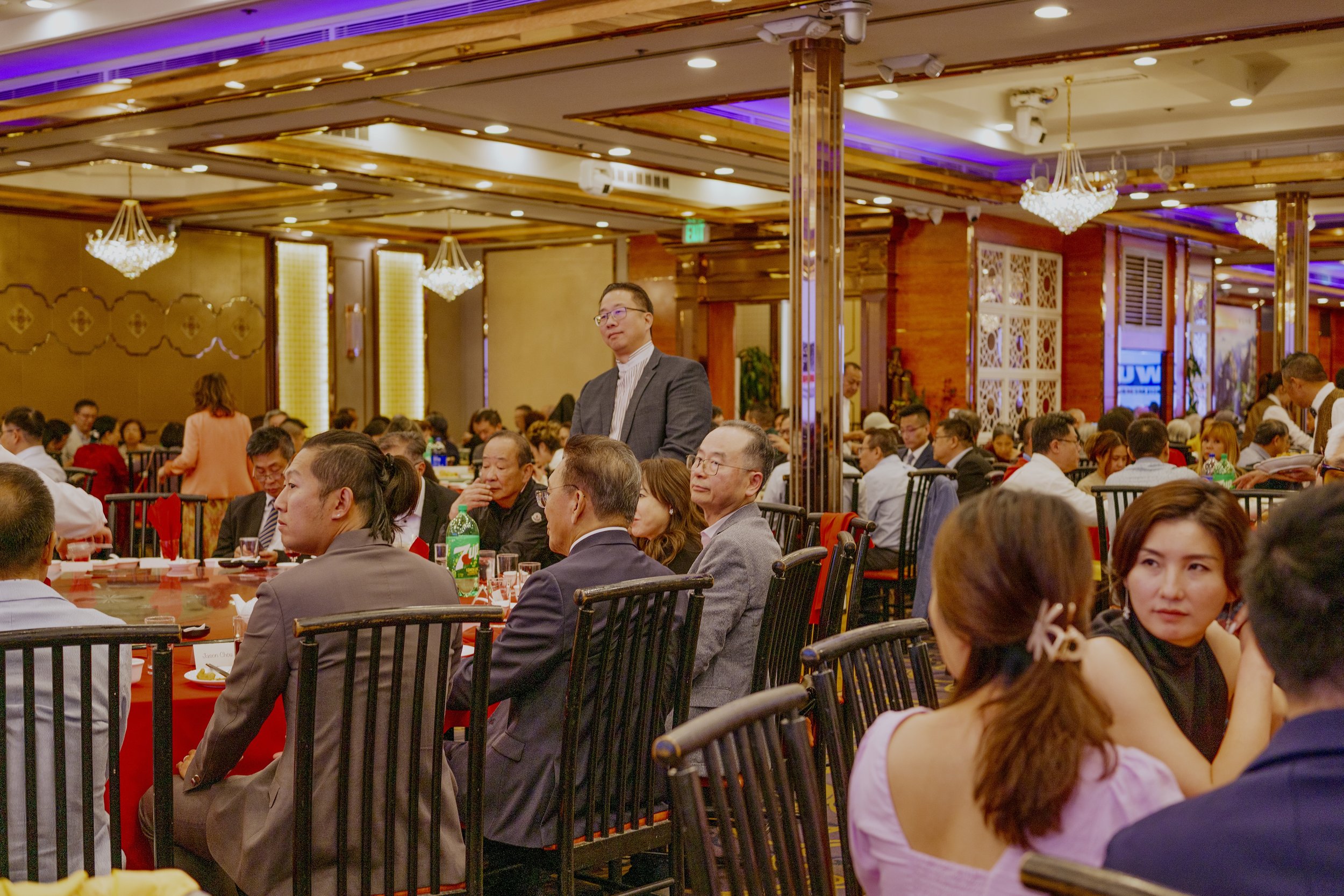 People sitting at tables in a large, elegant banquet hall with chandeliers and ornate wood and gold accents, attending a formal event or dinner.