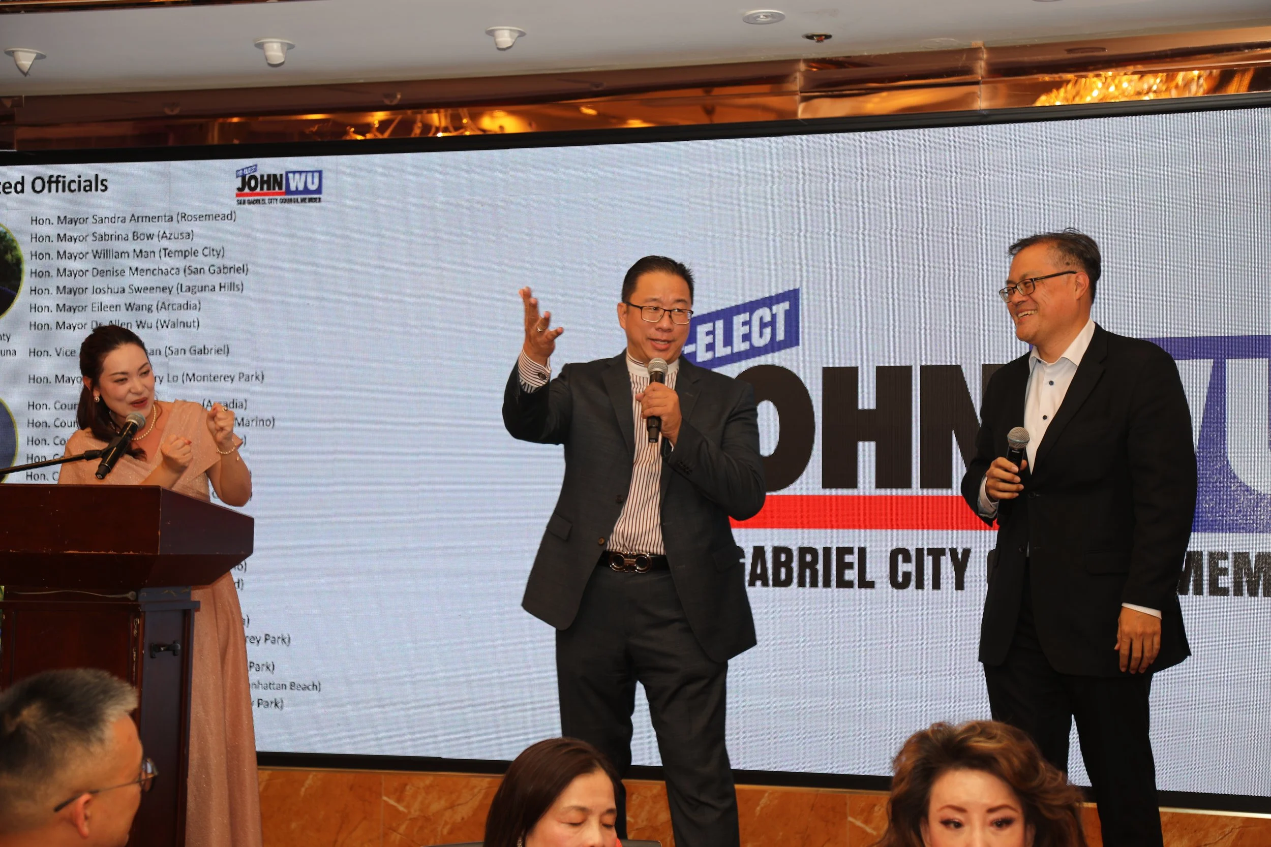 Three people on stage at an event, two men and one woman, with a large screen behind them displaying text. The man in the center is speaking into a microphone, the woman is at a podium, and the third man is smiling while holding a microphone.