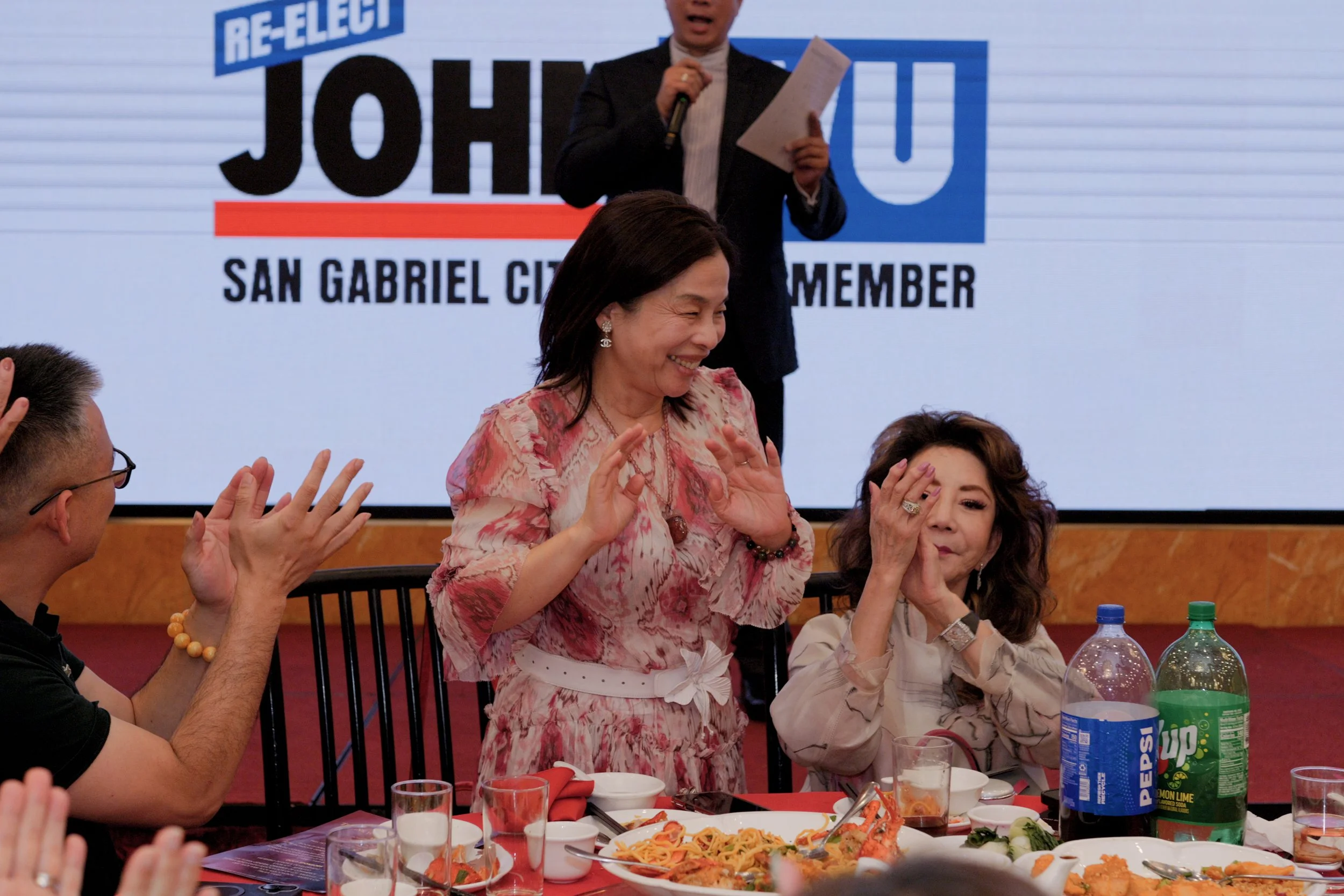 A group of people at a banquet table with food and drinks, celebrating, with a woman standing and smiling. A large screen in the background displays a political campaign message for John, a candidate for San Gabriel City Council.