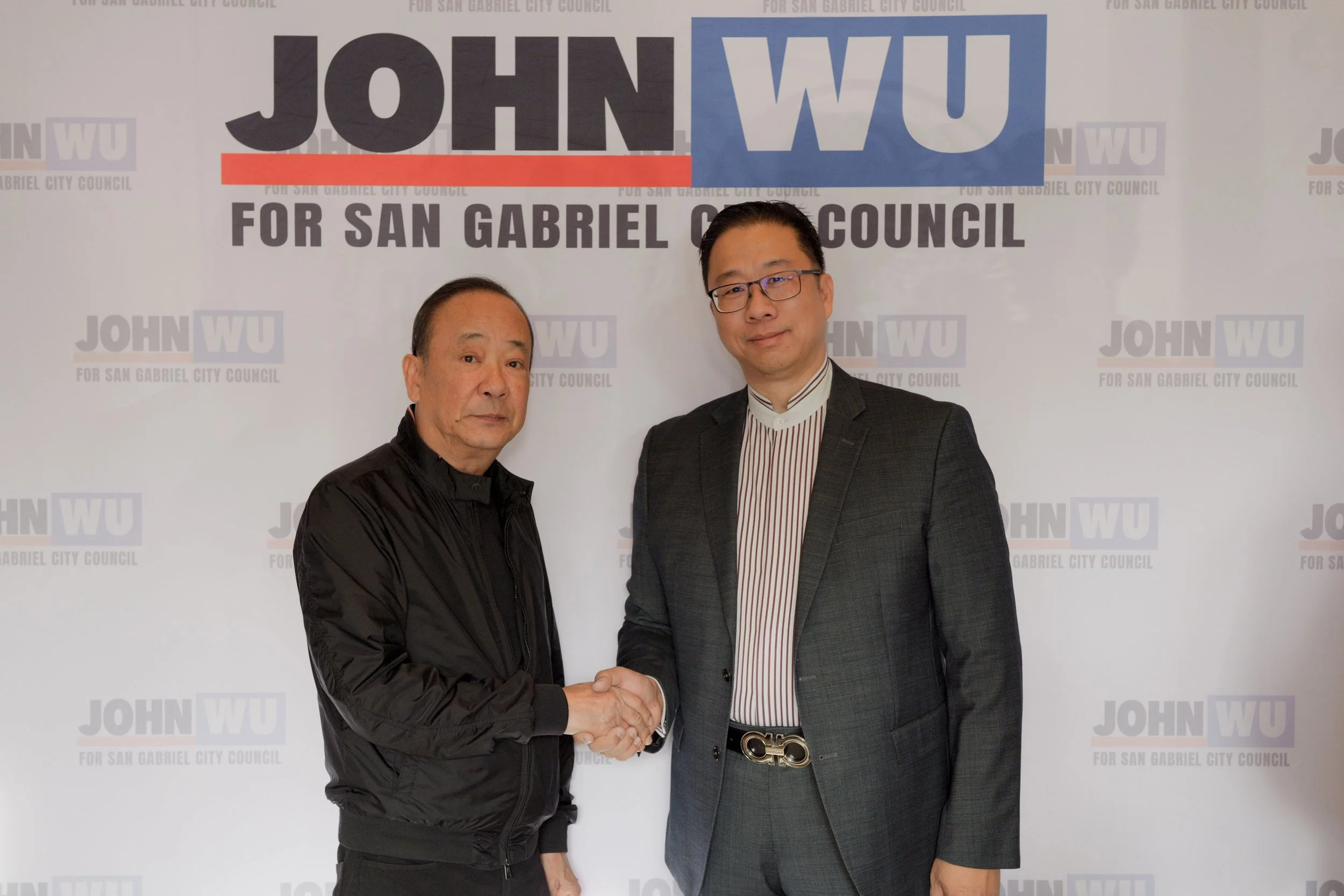 Two men shaking hands and posing for a photo in front of a backdrop that reads 'John Wu for San Gabriel City Council.' One man is wearing a black jacket and the other is wearing a dark suit with a striped shirt and Gucci belt.