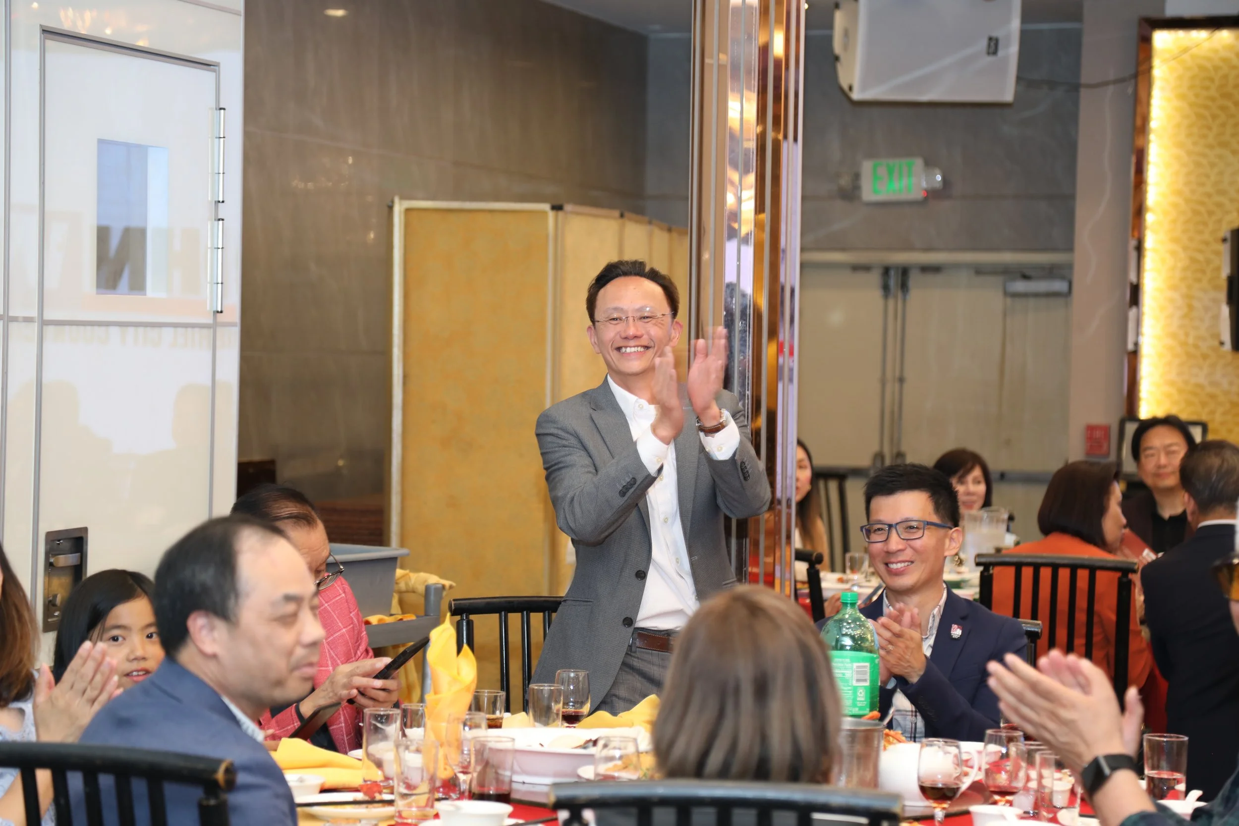 A man in a gray suit is clapping and smiling at a gathering or celebration, surrounded by seated individuals at a banquet table with yellow napkins and drinks.
