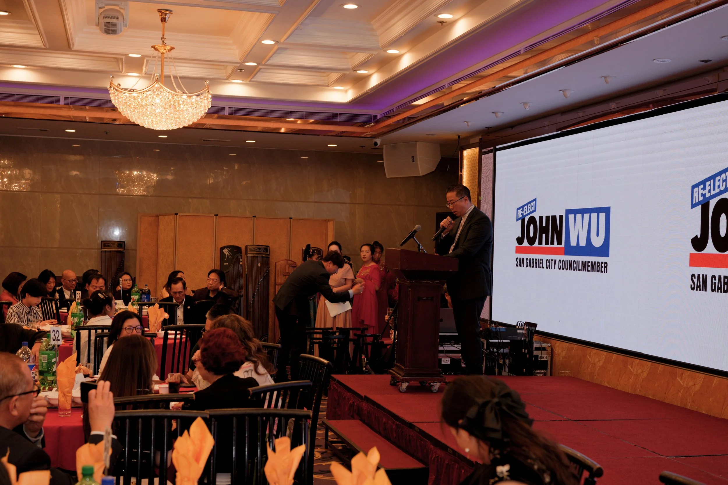 A speaker stands at a podium on a stage at a formal event, addressing a seated audience in a banquet hall decorated with chandeliers and ceiling lights. There is a large screen displaying campaign information for John Wu, a San Gabriel City Council M