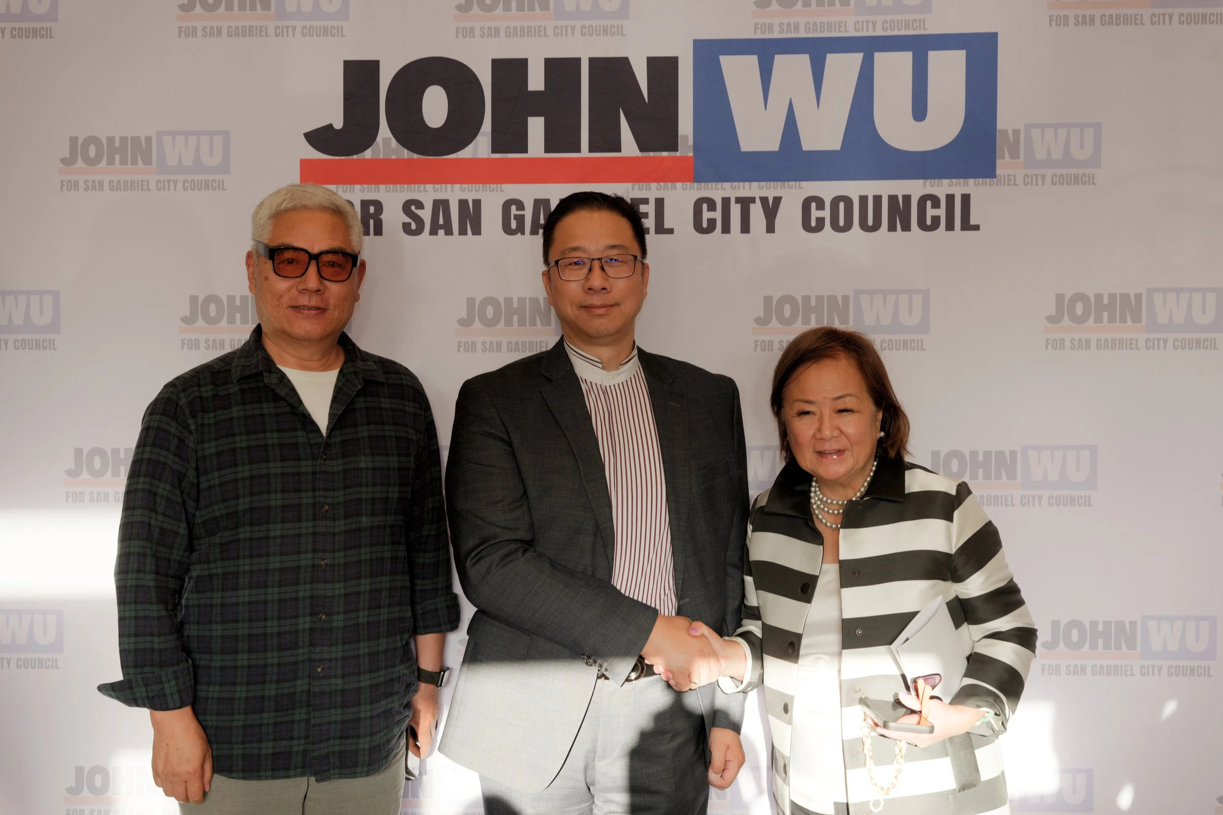 Three individuals on a stage or at an event, standing in front of a backdrop with political campaign text. The backdrop features the name 'John Wu' and mentions San Gabriel City Council. One person is greeting or shaking hands with another, while the