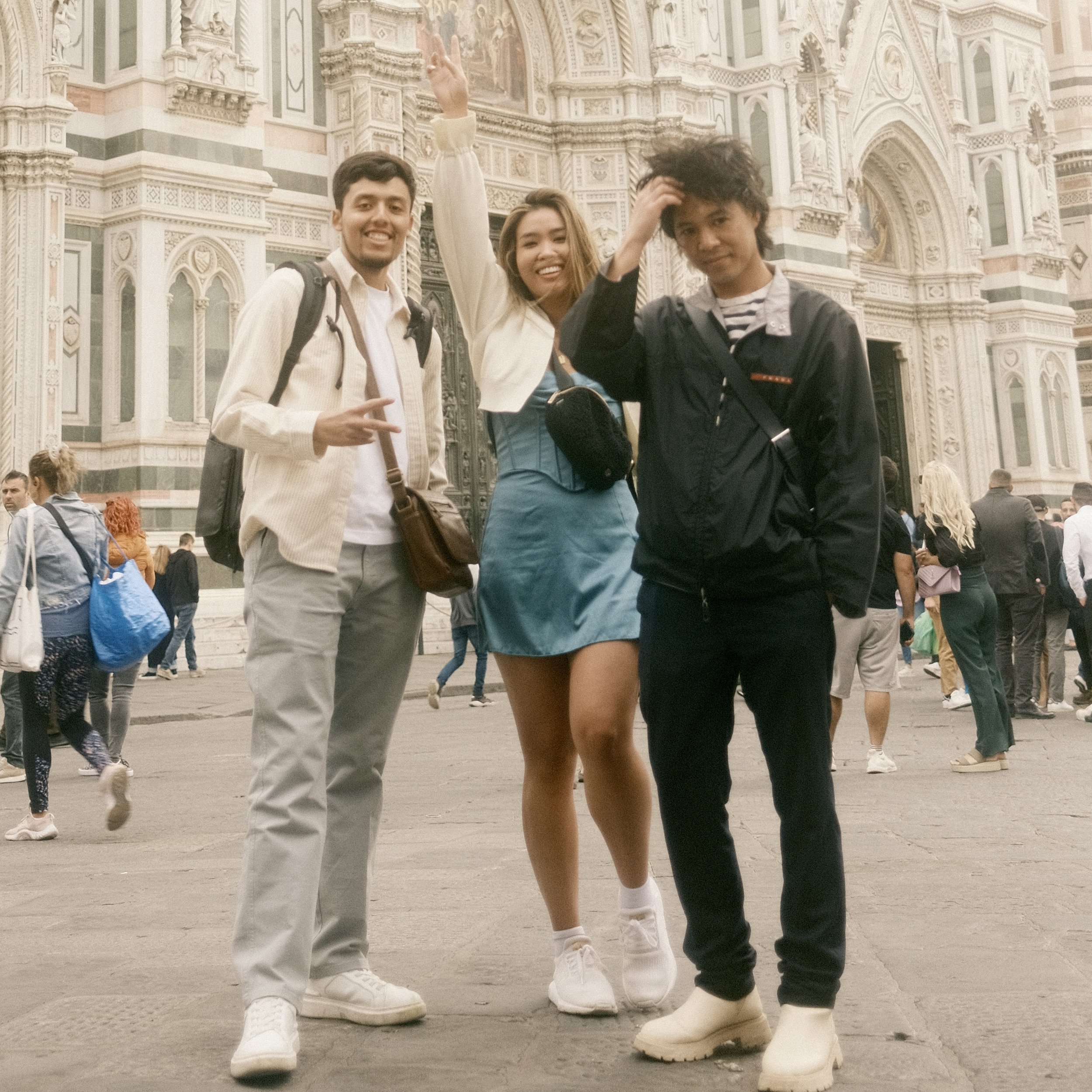 Three young people standing in front of a historical building, smiling and posing for a photo. The person on the left is a man with short dark hair, wearing a light-colored shirt and carrying a backpack. The person in the middle is a woman with long blond hair, wearing a white jacket and a blue skirt. The person on the right is a man with curly dark hair, wearing a black jacket and striped shirt. The background shows a crowd of people and intricate architecture on the building.