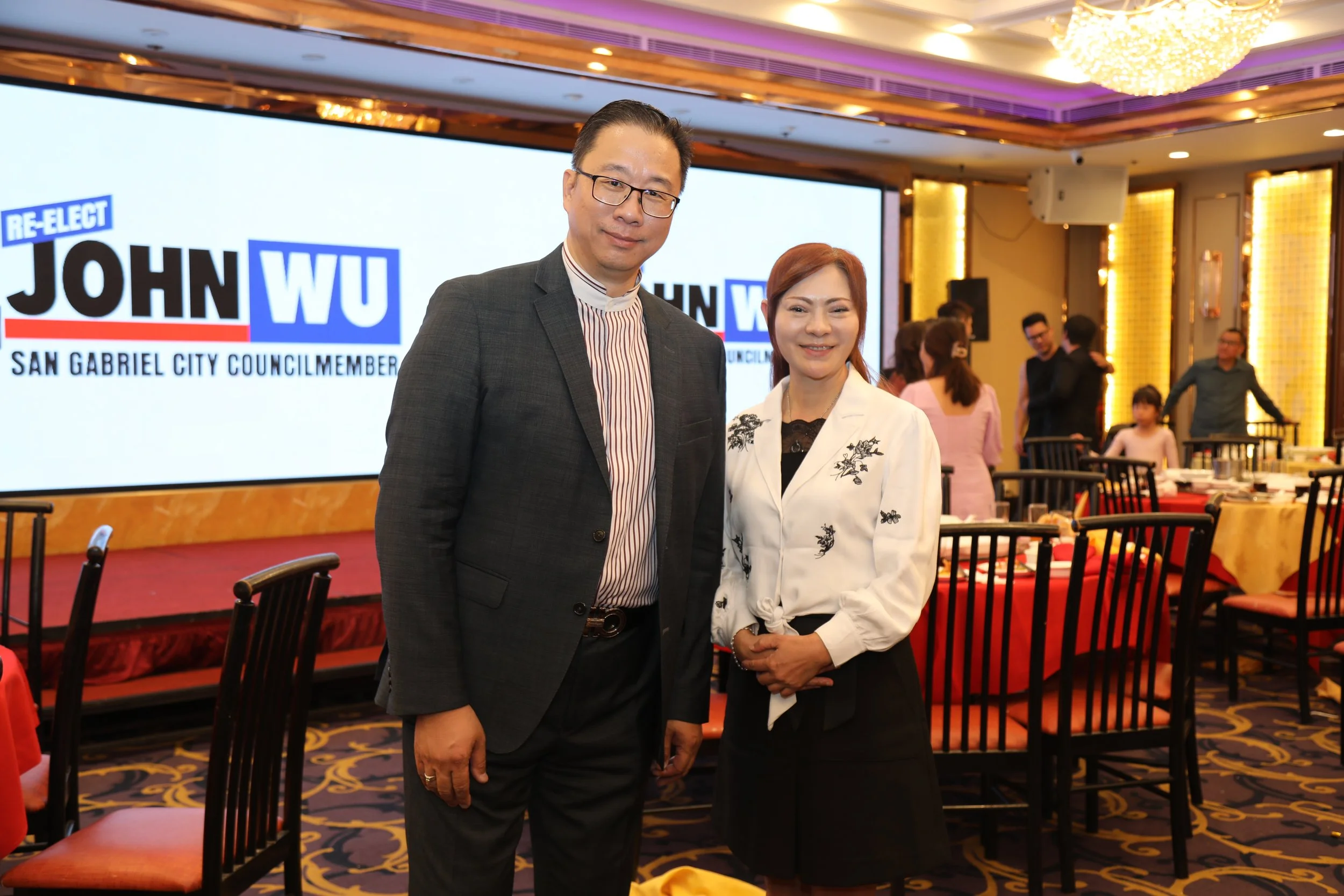 Two people, a man and a woman, standing together in a banquet hall or conference room. The background features a large screen displaying the name 'John WU' and the title 'San Gabriel City Councilmember'. The room is decorated with warm lighting, chan