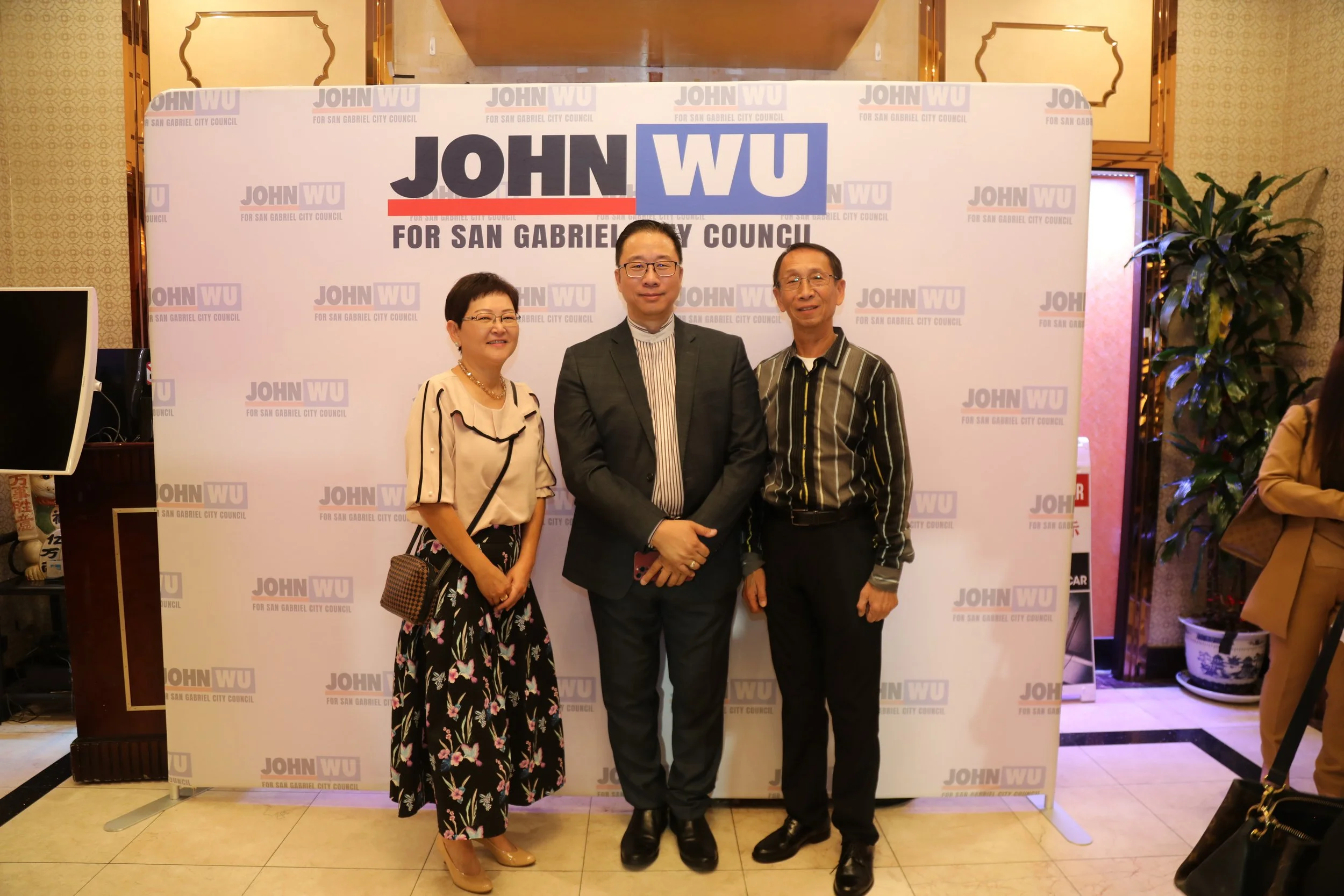 Three people standing in front of a campaign backdrop that reads 'John WU for San Gabriel City Council.' The woman on the left is wearing a beige blouse with black accents and a floral skirt. The man in the center is wearing glasses and a dark suit w