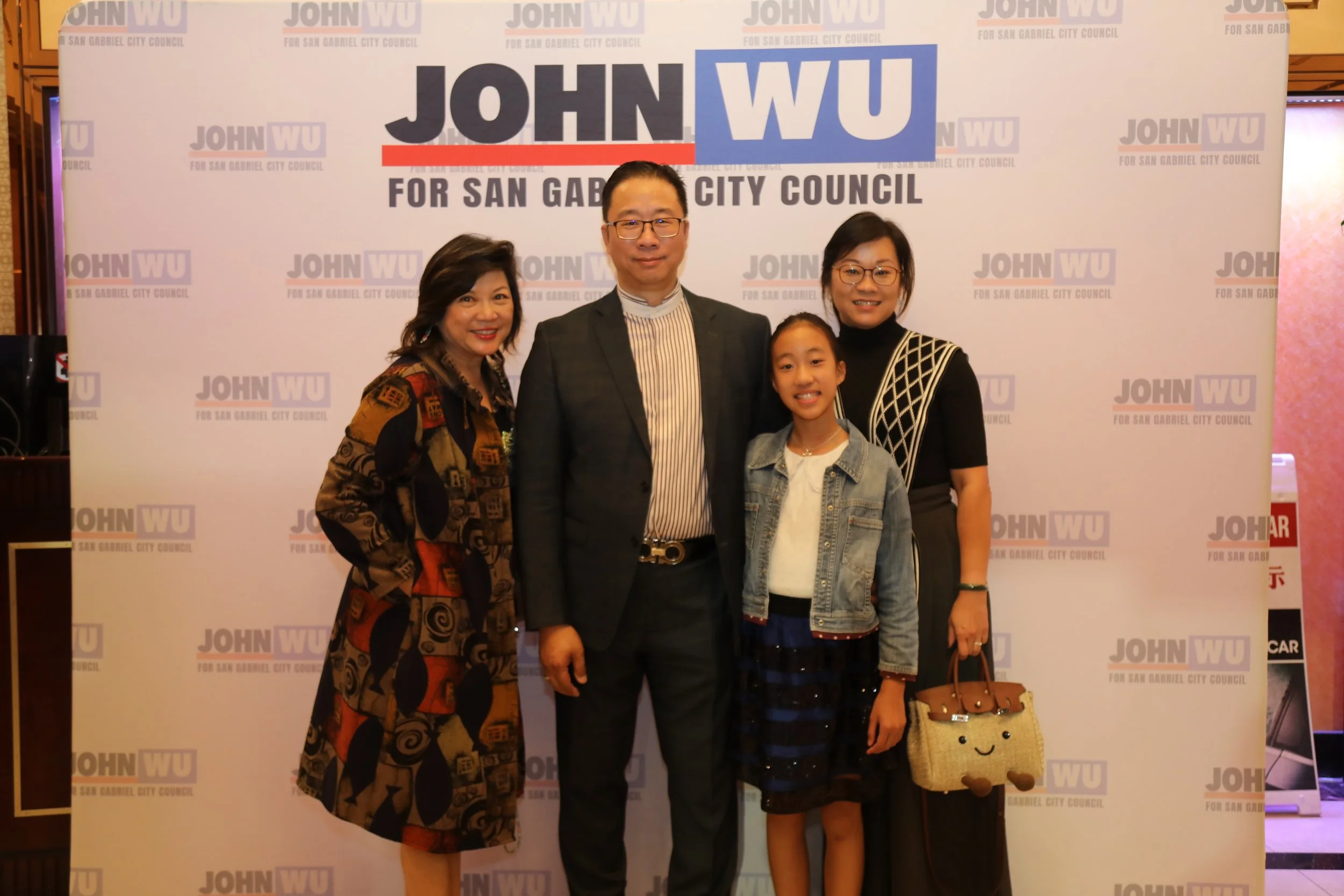 Four individuals standing together in front of a backdrop with the text 'JOHN WU FOR SAN GABRIEL CITY COUNCIL'. The group includes two women, one man, and a young girl, all smiling and posing for the photo.