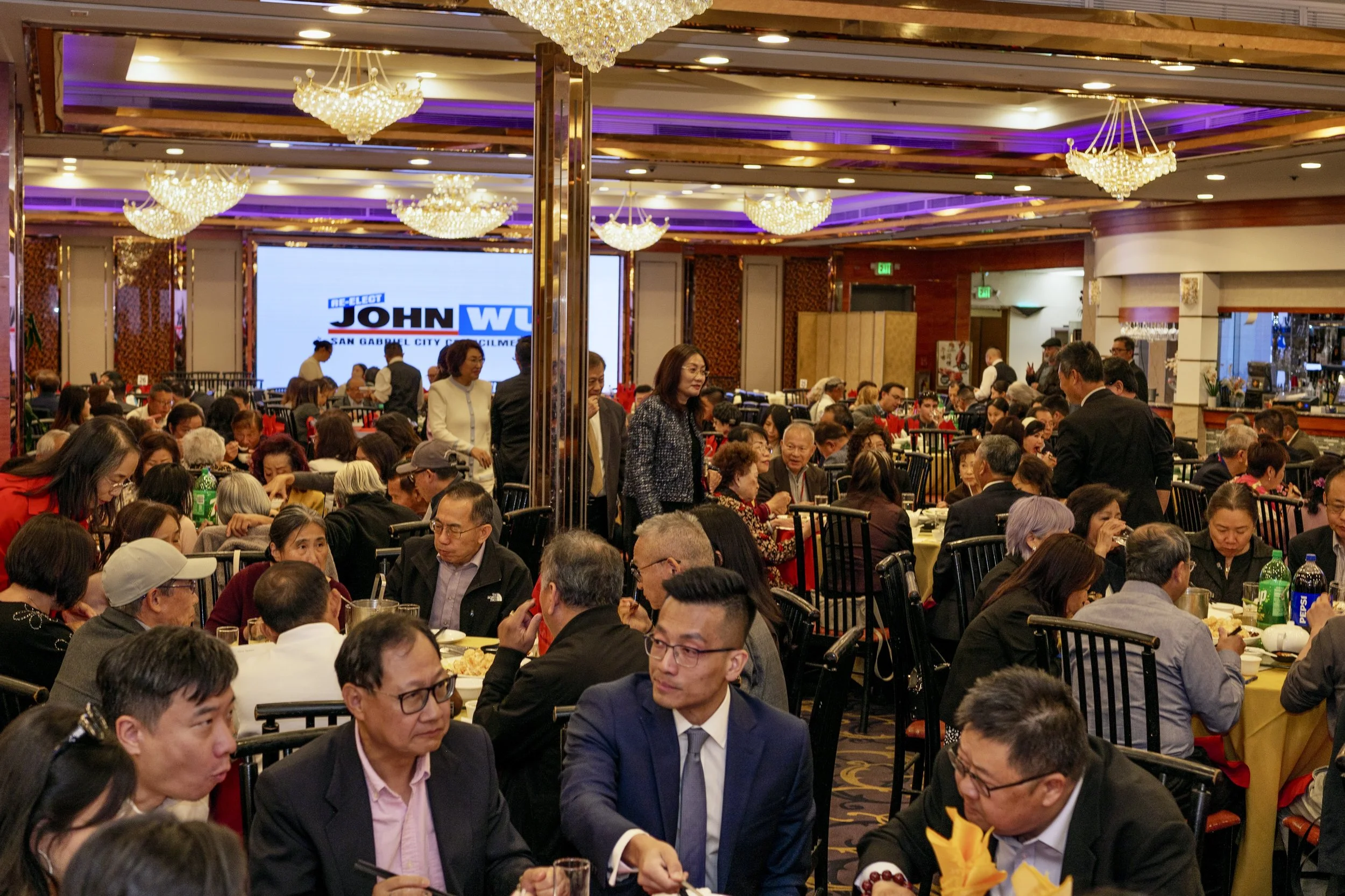 A large banquet hall filled with people sitting at tables, enjoying a meal, with elegant chandeliers hanging from the ceiling, and a large screen displaying a campaign logo in the background.