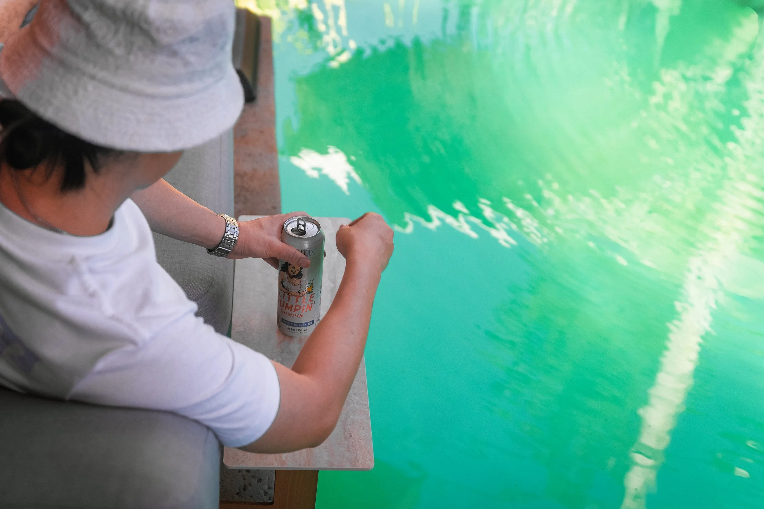 Overhead view of a person in a bucket hat opening a drink can on the chaise lounge side table above green pool water