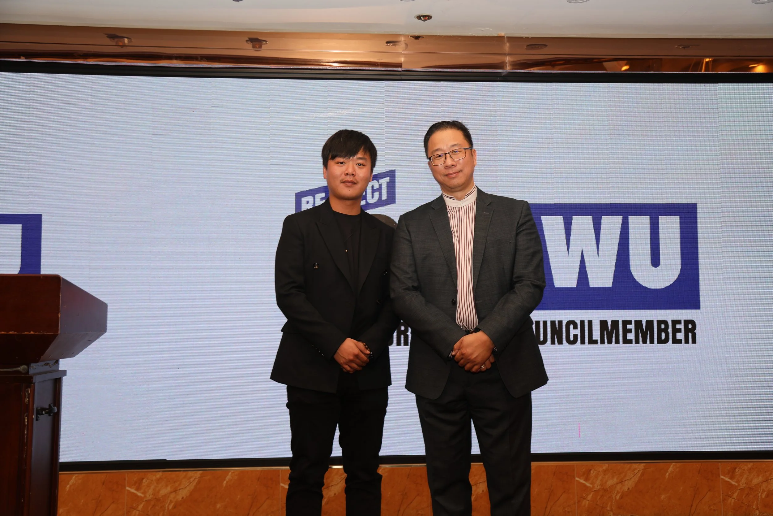 Two men in suits standing on a stage in front of a large screen that reads 'Respect' and 'Council Member'.