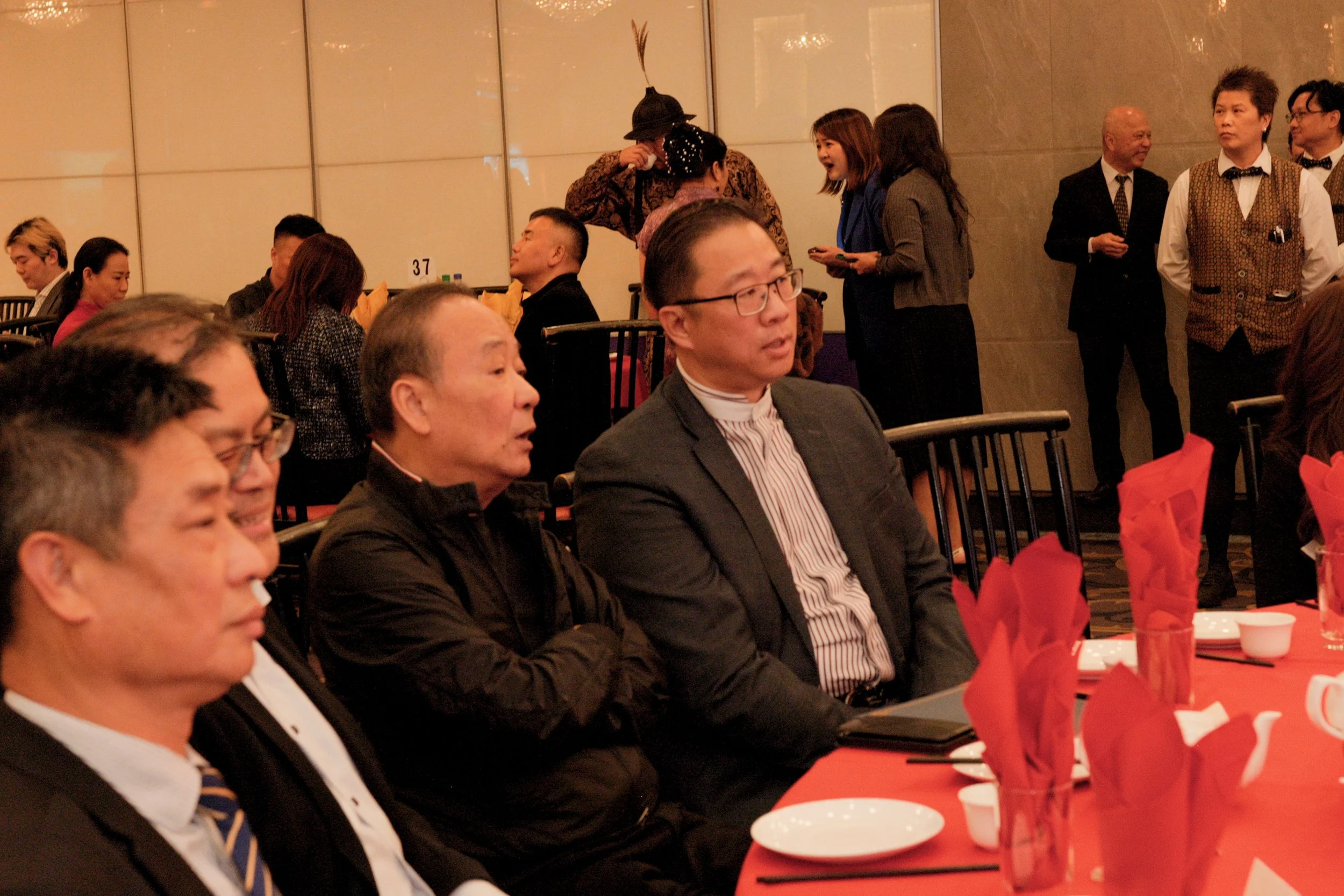 People seated at a banquet table decorated with red napkins, engaged in conversation at a formal event in a banquet hall.