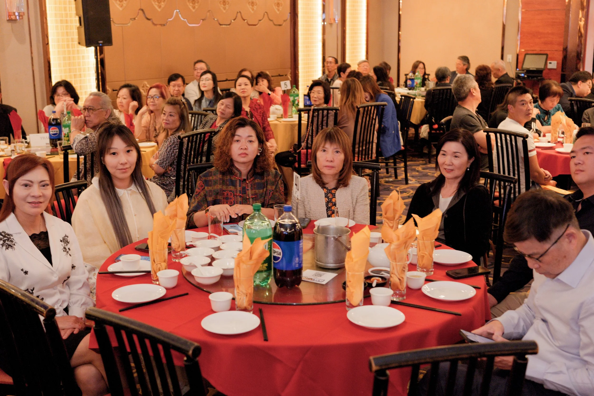 A group of Asian and Caucasian people seated around a banquet table at a social event, with some looking towards the front and others engaged in conversation. The table is set with orange napkins, white dishes, and various beverages including soda bo