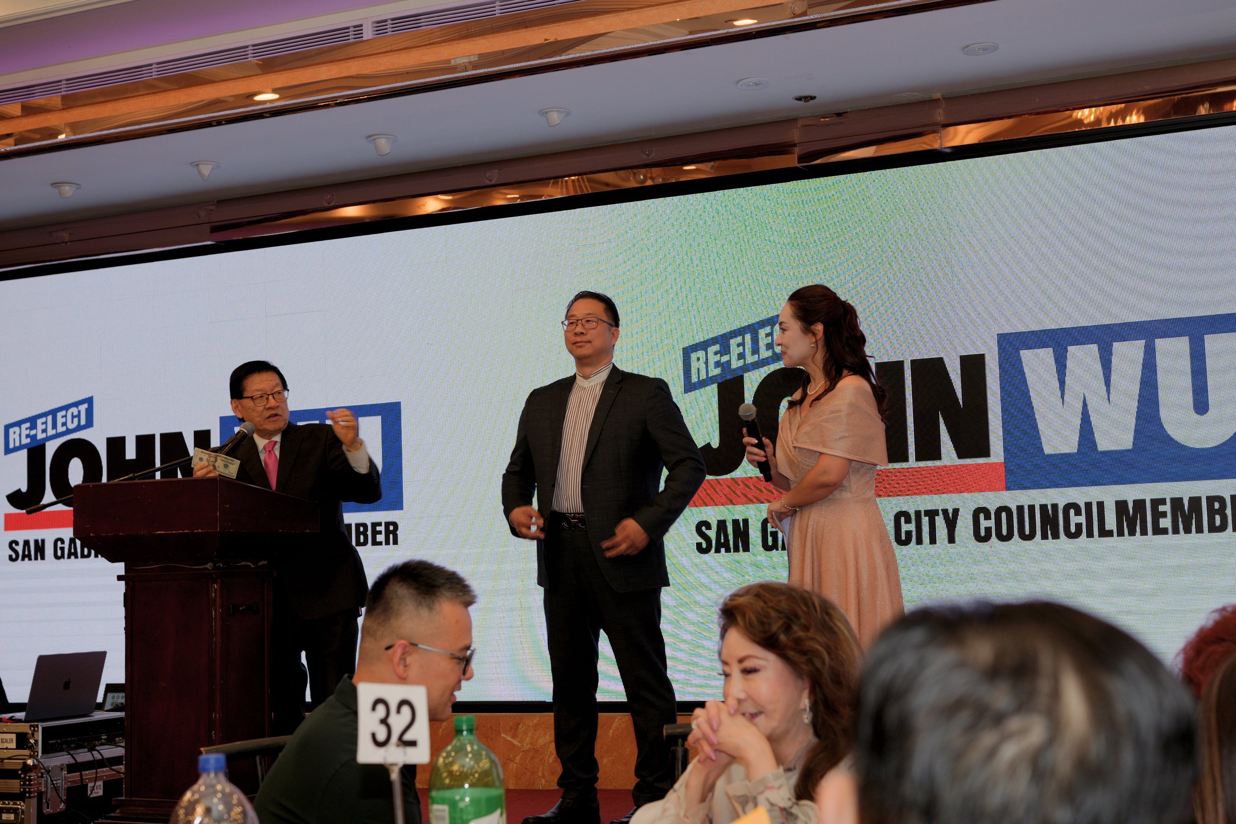 A formal event with three individuals on stage, a man in a suit and tie, a man in a suit, and a woman in a dress, standing in front of a large screen displaying the words 'Re-Elect John Wu' and 'San Gabriel City Council Member'. The man on the left i