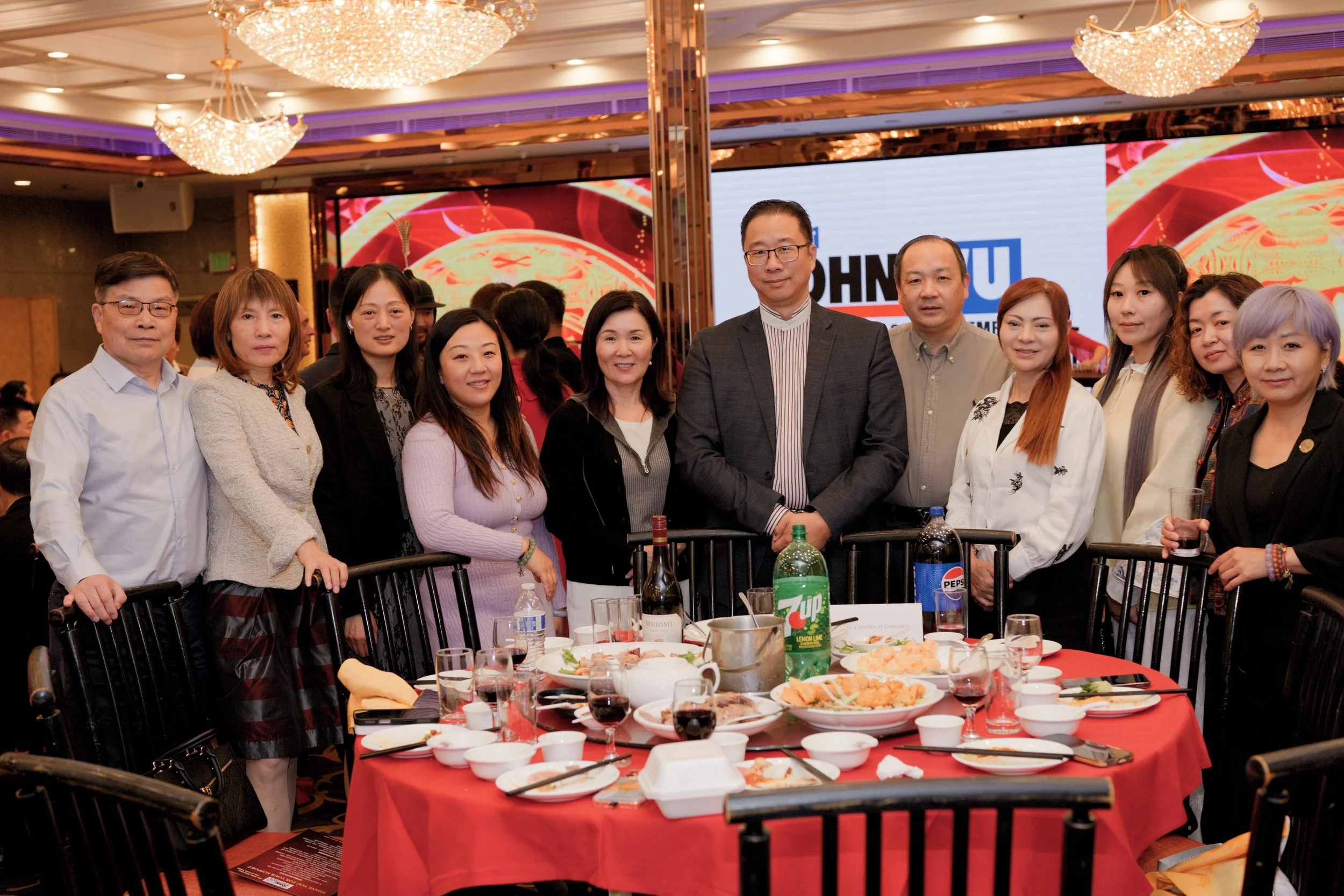 Group of people posing at a banquet table with food, drinks, and a large digital screen in the background showing a colorful design and the word 'JOHN'. The setting appears to be a formal event or celebration in a banquet hall with chandeliers.