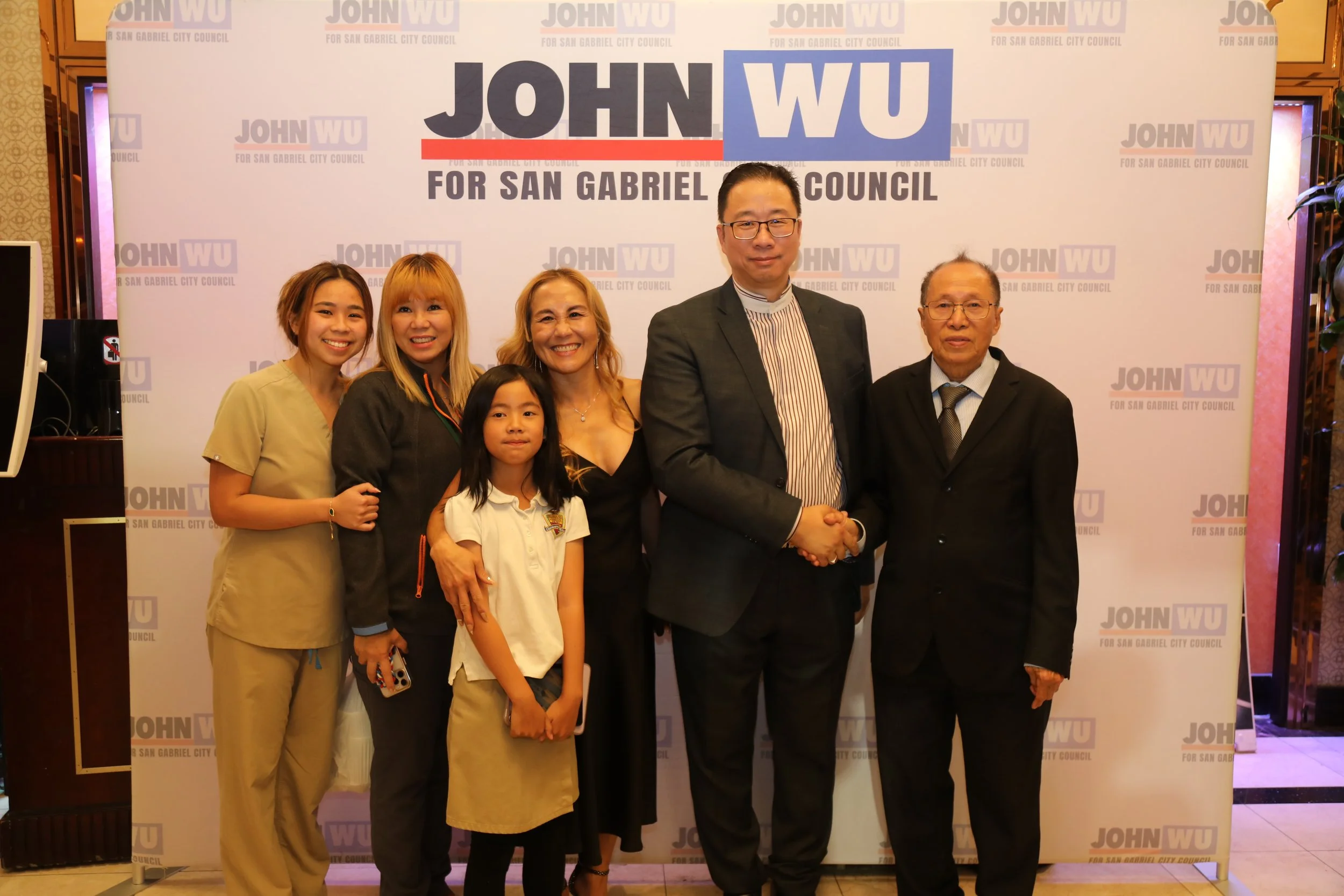 A group of six diverse people, including men, women, and a girl, standing in front of a campaign backdrop for John Wu running for San Gabriel City Council. They are smiling and appear to be at a political event or campaign gathering.
