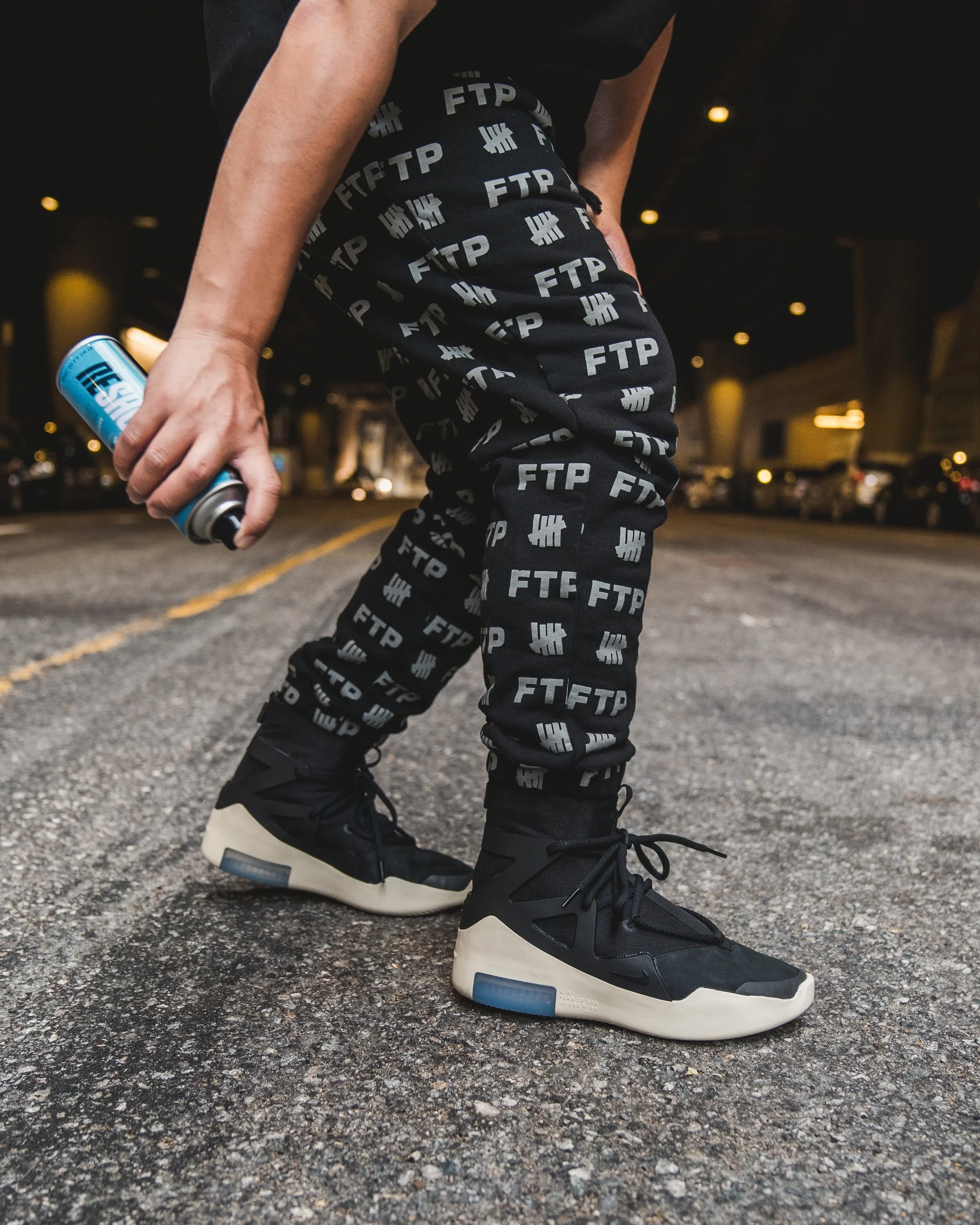 Person wearing black pants with 'FTP' logos, black and white sneakers, standing on a city street at night, holding a spray can.