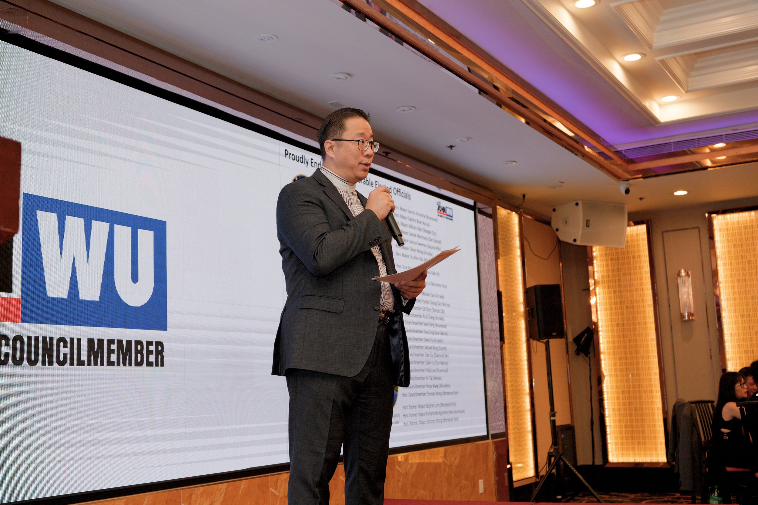 A man in a gray suit, glasses, and a striped shirt speaking into a microphone during a presentation at a conference or meeting, with a large screen behind him displaying the logo of the Washington University in St. Louis and text about election offic